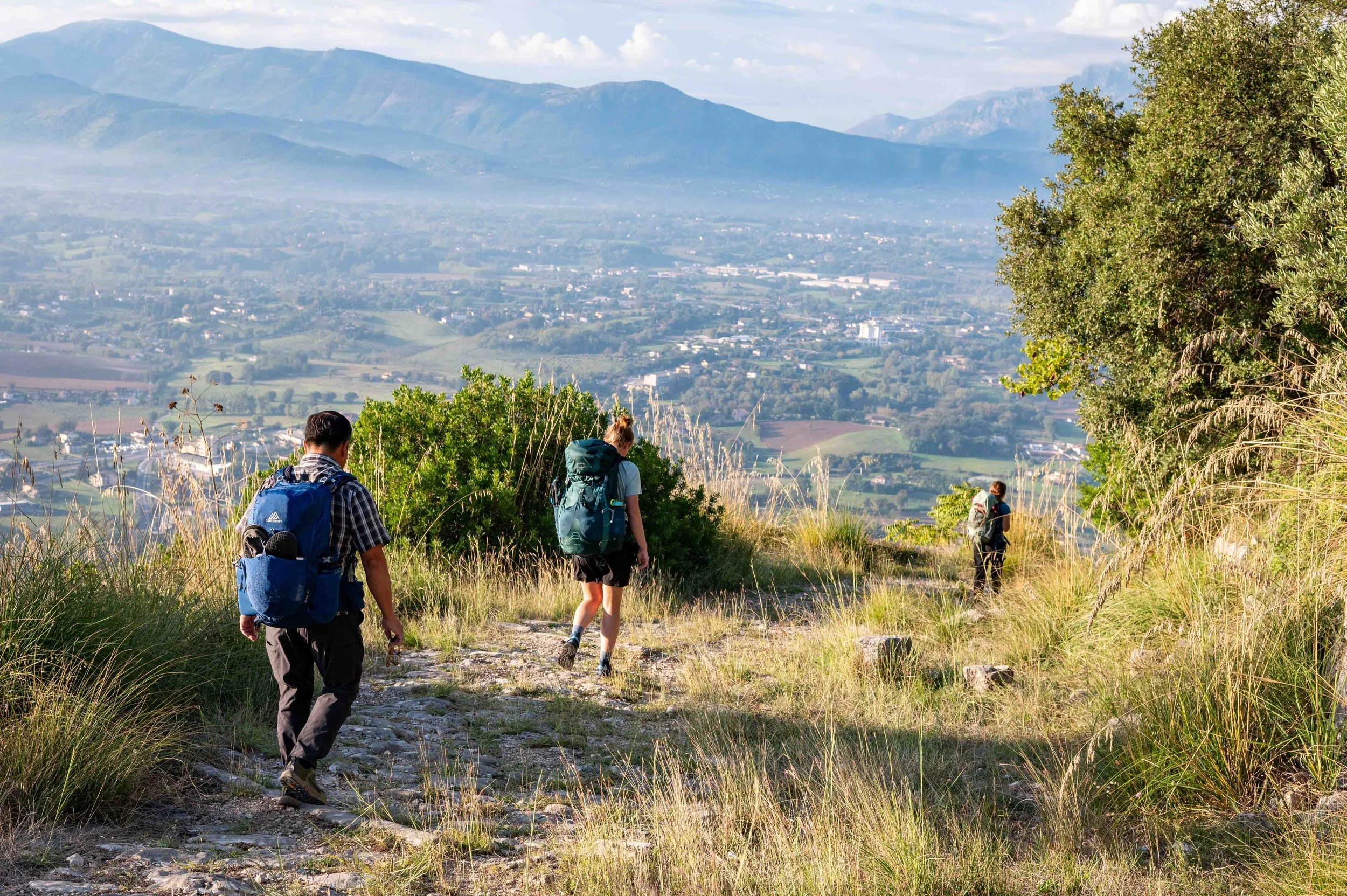 Three hikers with backpacks walking down a grassy trail on a mountainside, overlooking a valley with mountains in the background.