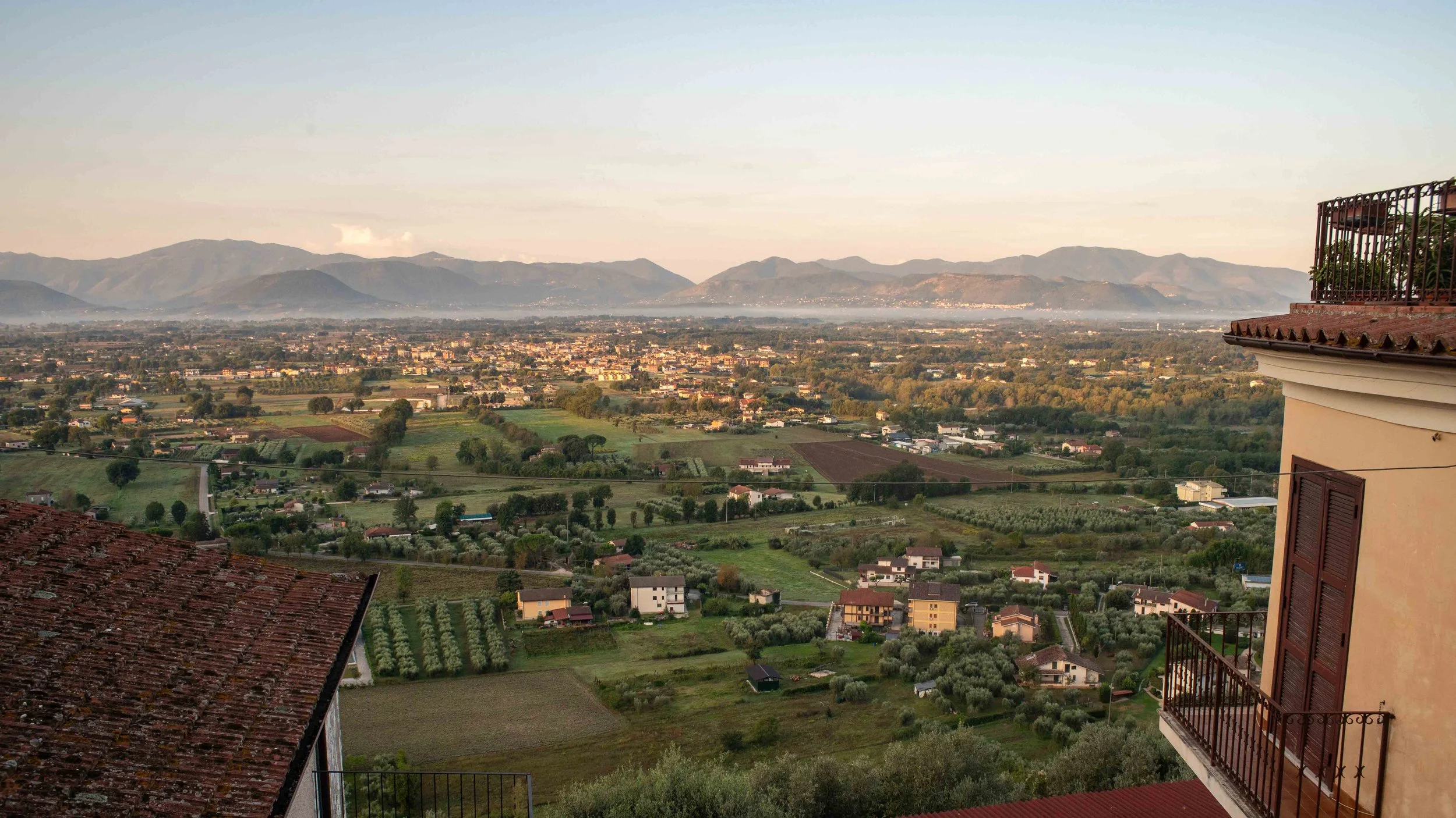 A scenic view of a valley with rolling green fields, scattered houses, and distant mountains at sunset.