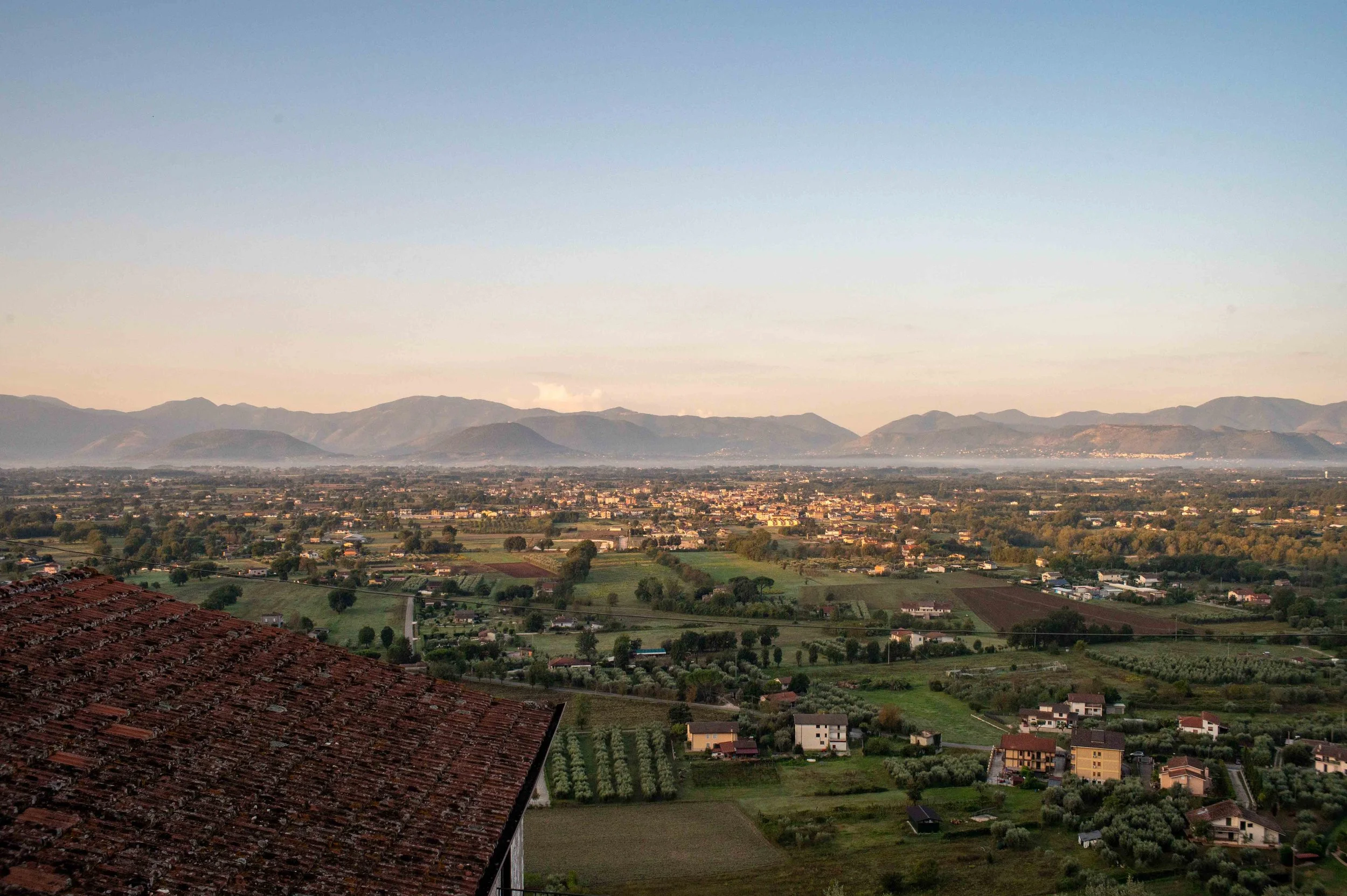 A wide view of a rural landscape with houses, green fields, and mountains in the distance under a clear sky at sunset.
