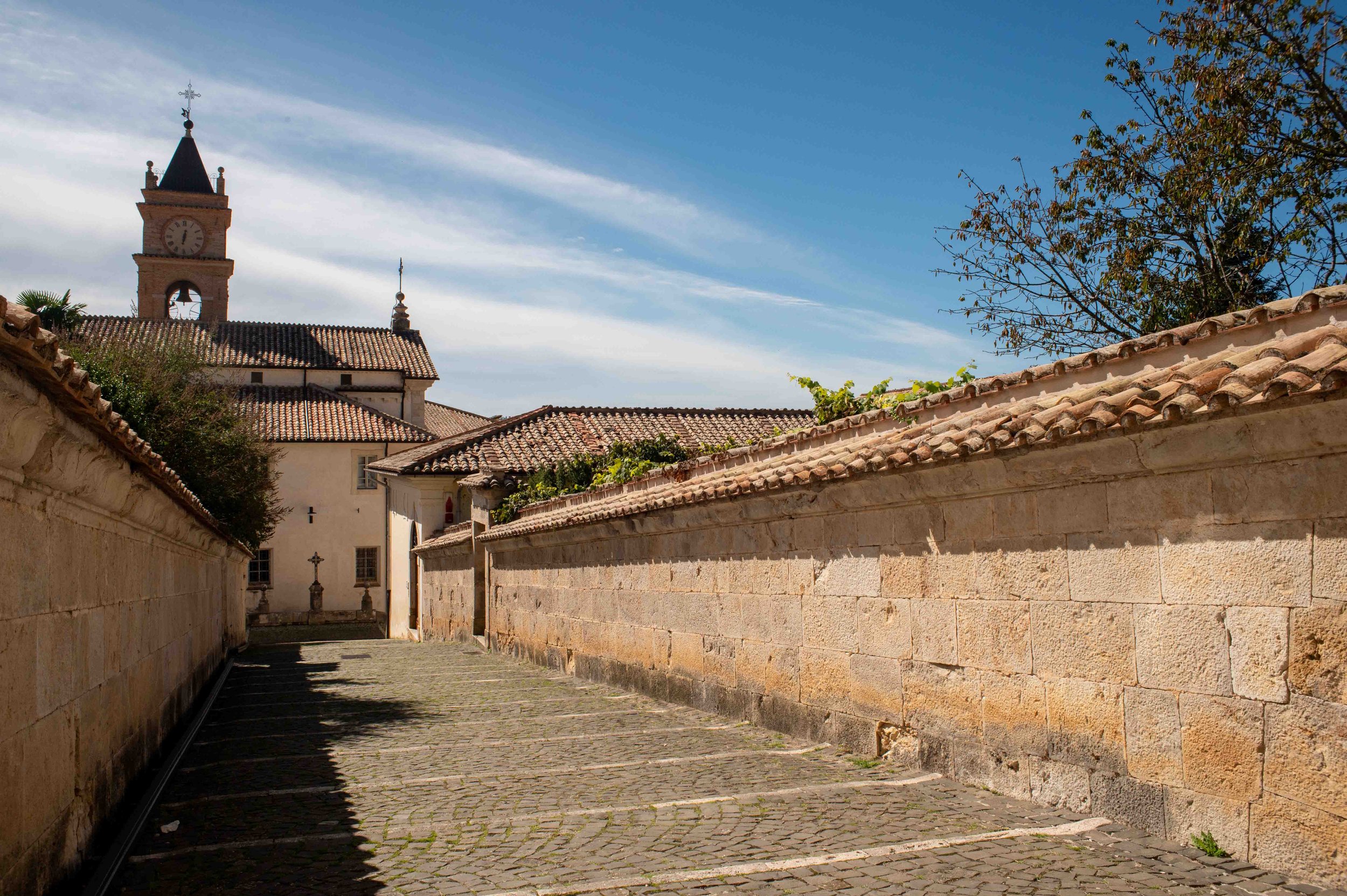 A narrow cobblestone street with stone walls on each side, leading to a church with a bell tower and a clock, under a partly cloudy blue sky with a tree on the right.