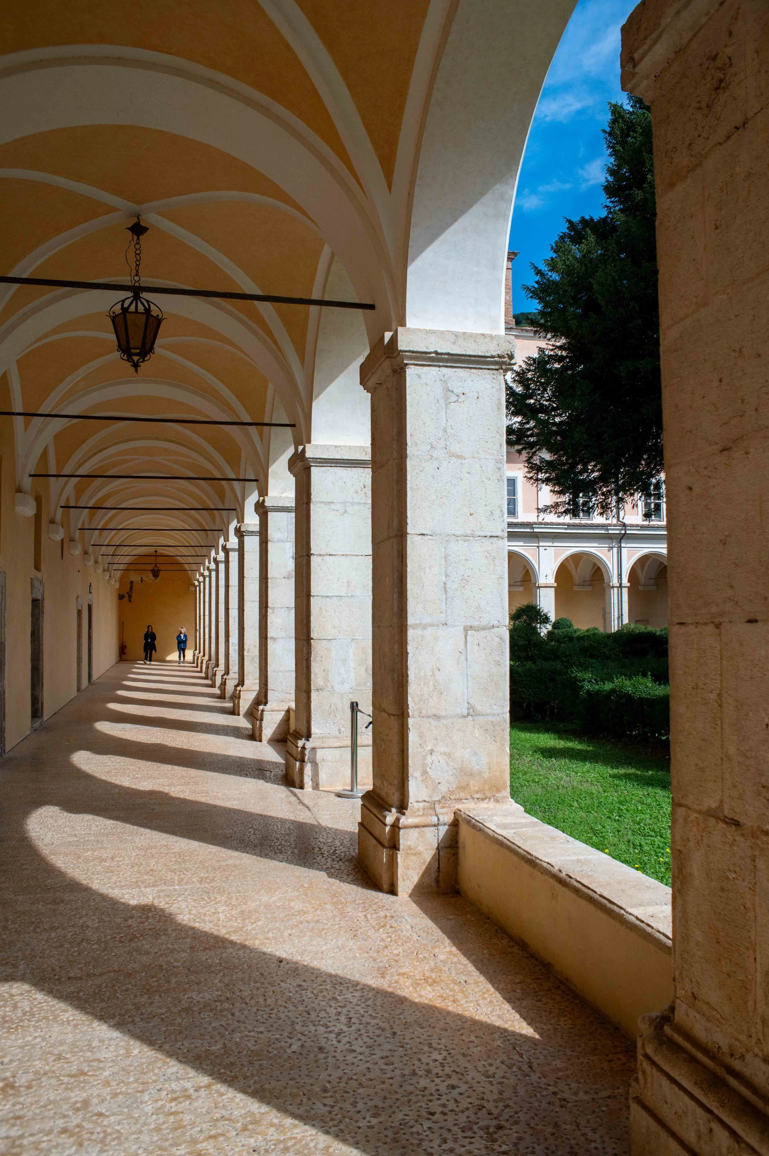A covered arched walkway with stone columns, hanging lanterns, and shadows cast on the ground, overlooking a grassy area with trees and a building in the background.