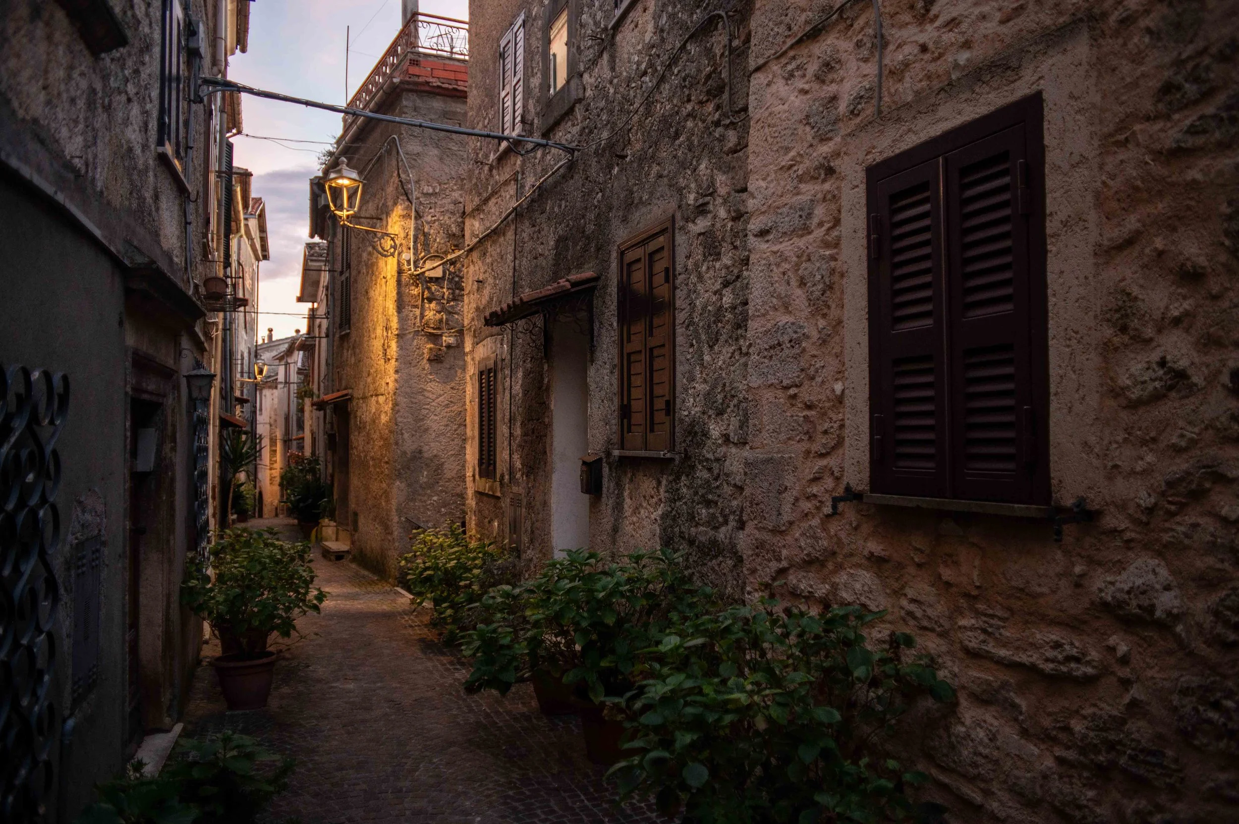 Narrow cobblestone alleyway in an old European city during dusk, with stone buildings on either side, small potted plants, and warm street lighting.