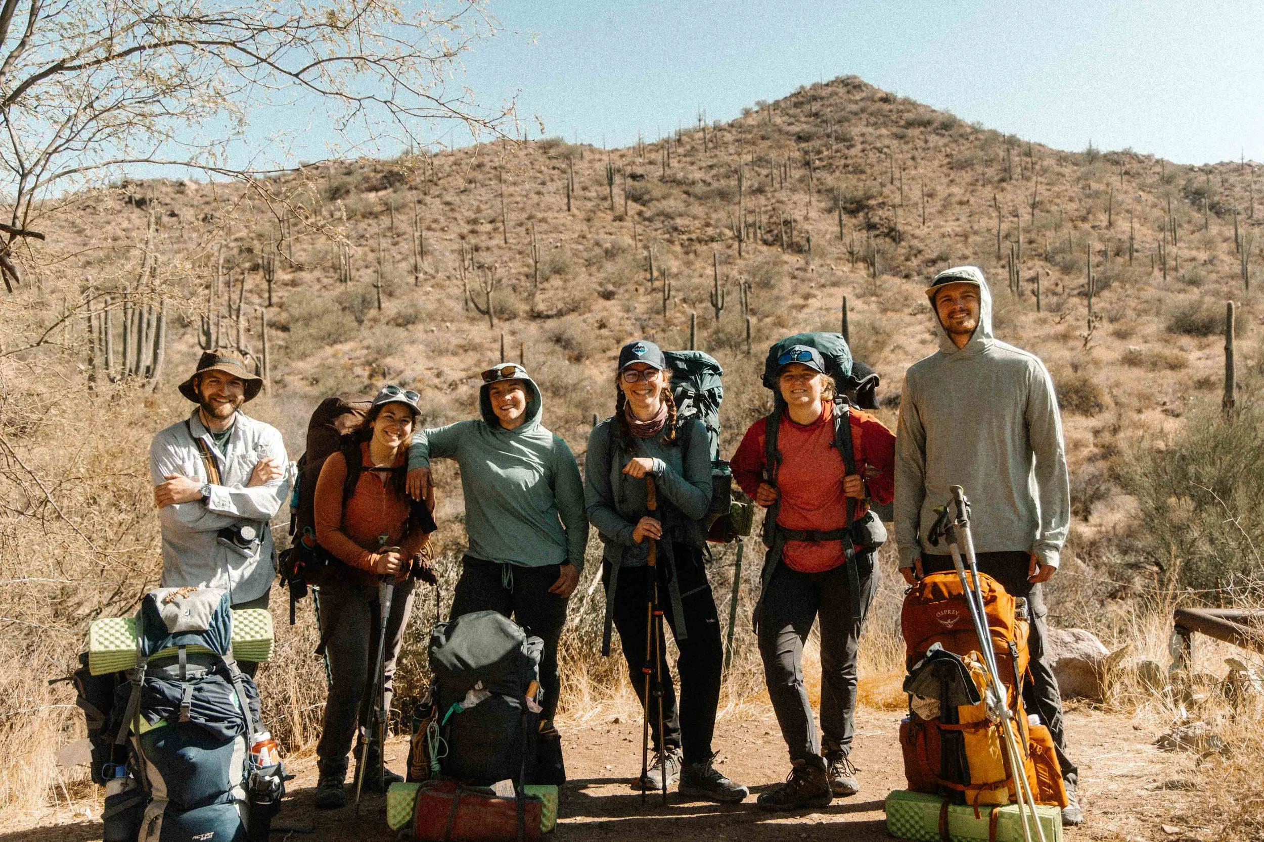 A group of six people with backpacks and hiking gear standing on a dirt trail in a desert landscape with cactus and dry shrubs, smiling at the camera.