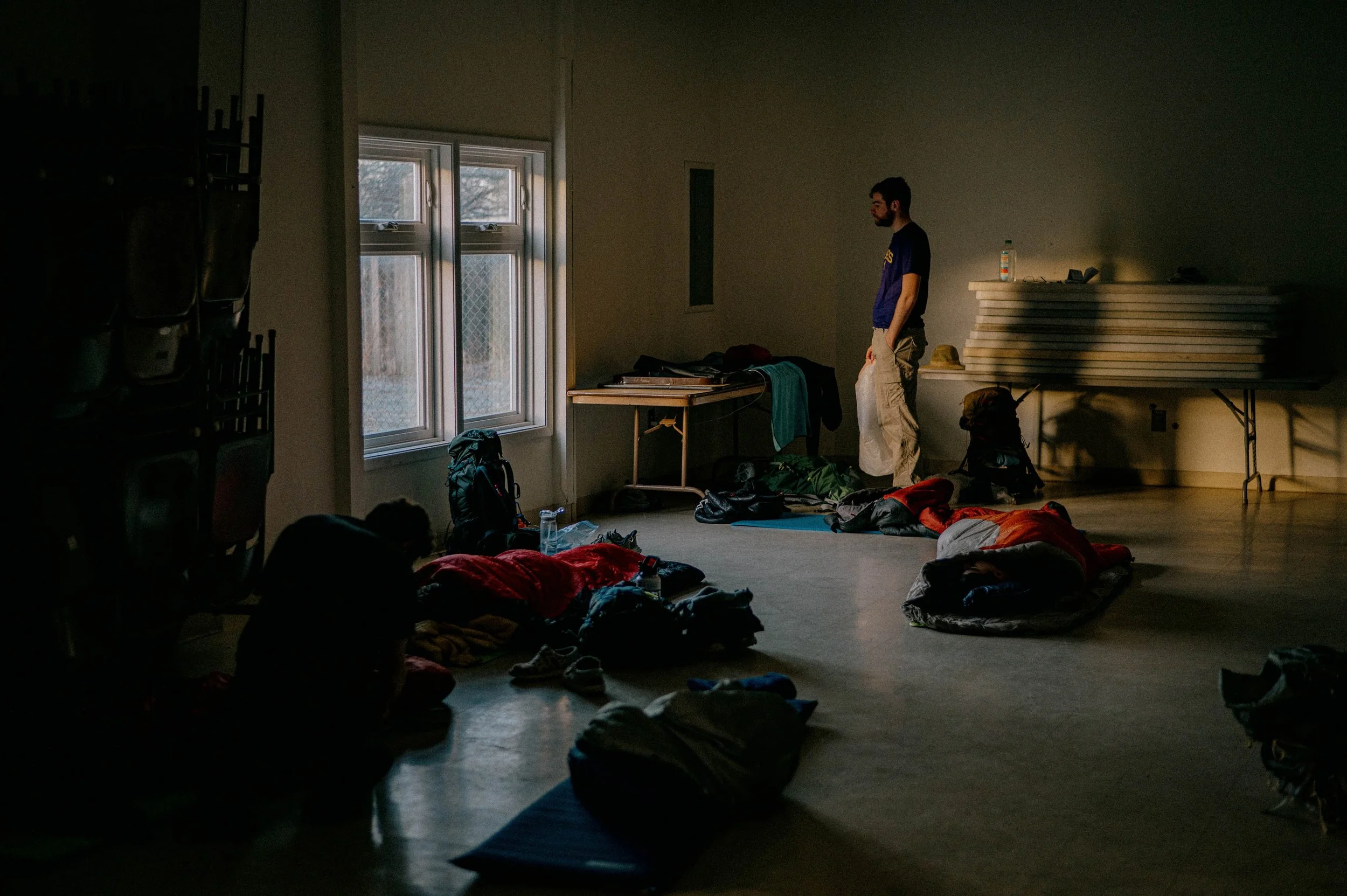 A dimly lit room with two people on the floor and one person standing near a window, surrounded by backpacks, sleeping bags, and supplies.