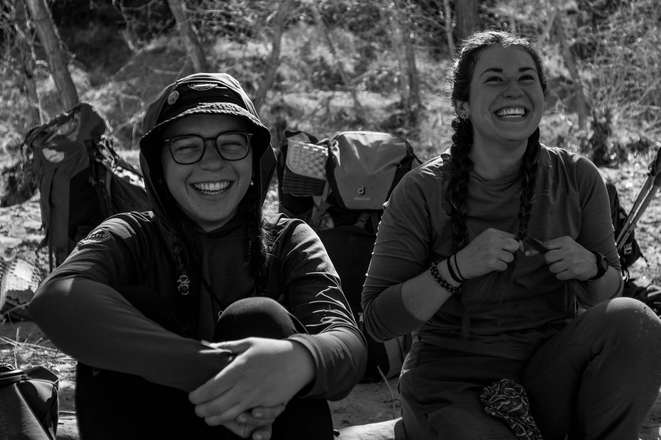 Two women sitting outdoors, smiling and laughing, surrounded by backpacks and outdoor gear, with trees in the background.