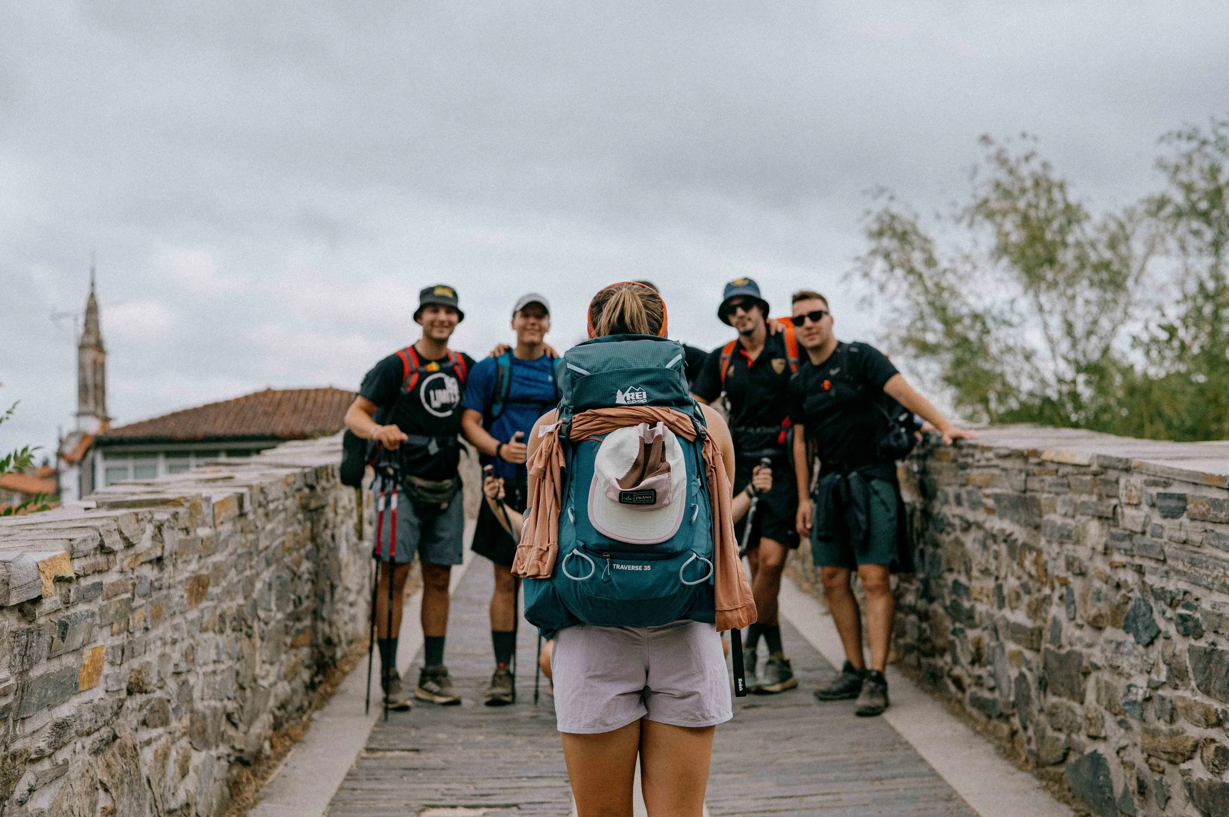 A woman with a large teal hiking backpack standing on a stone bridge facing five men, with a town and church steeple in the background under a cloudy sky.