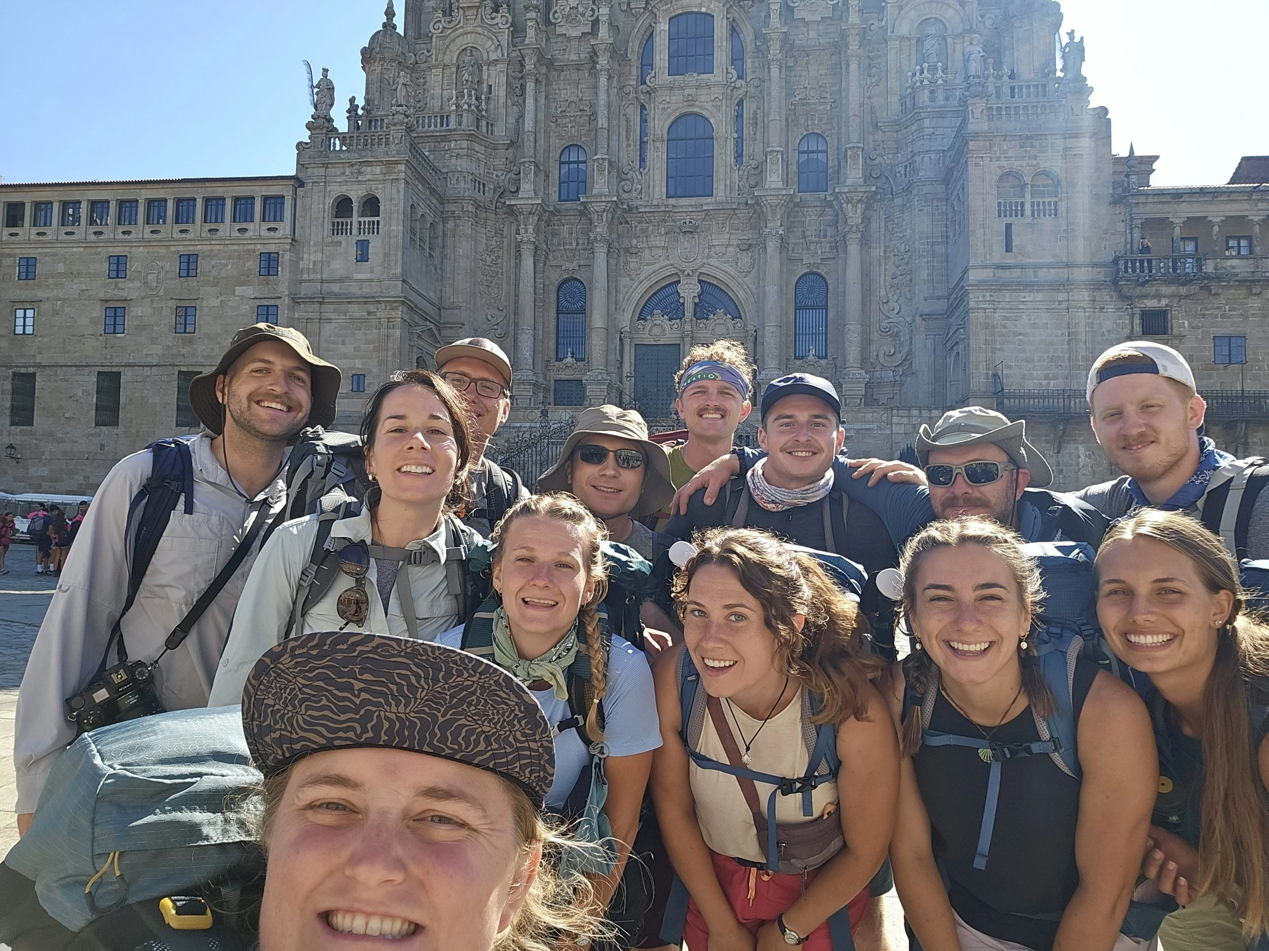 A group of smiling hikers, both men and women, posing in front of a historic, ornate building with detailed architecture, likely a cathedral, on a sunny day.