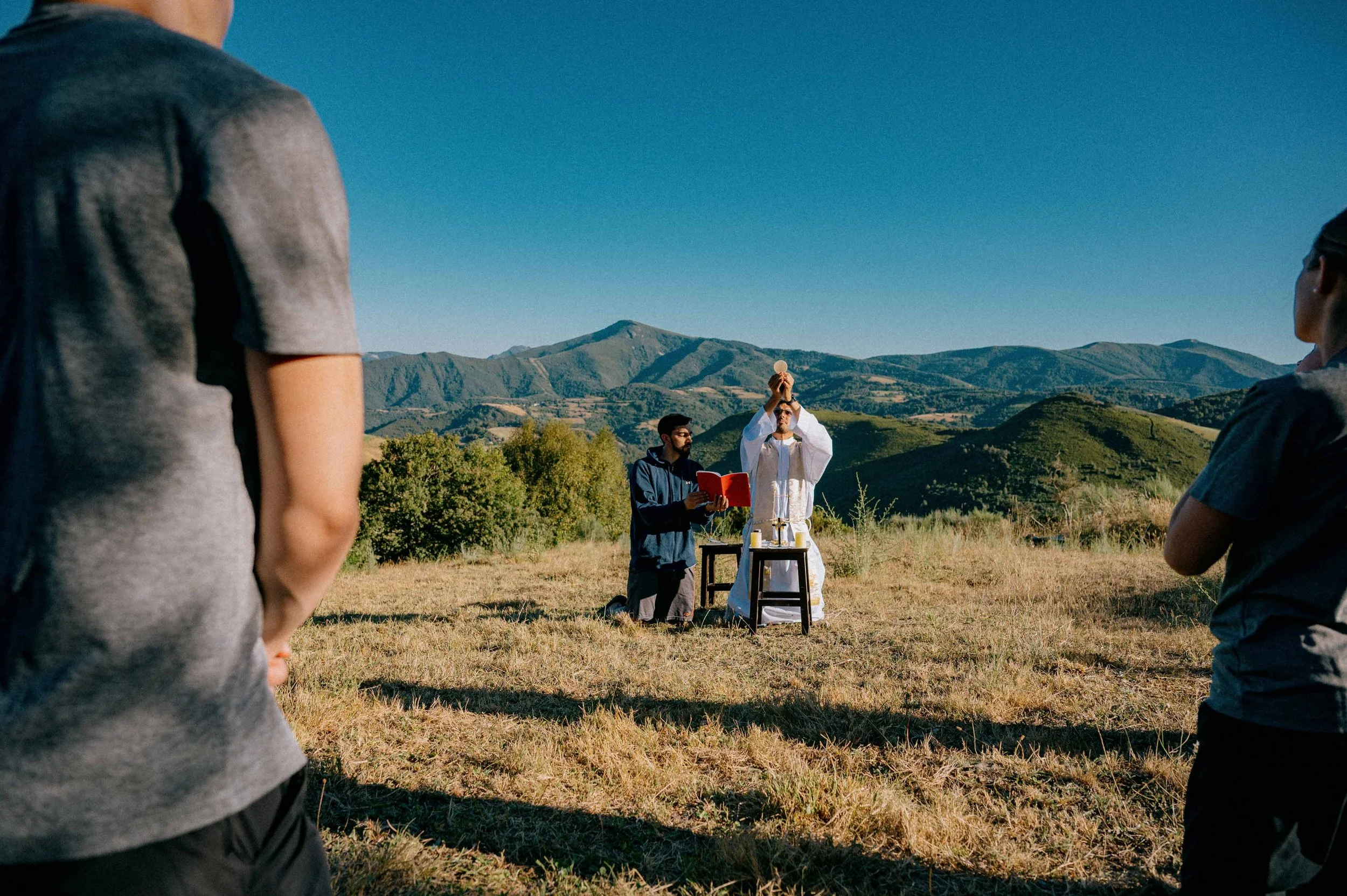 A religious ceremony taking place outdoors in a field with a mountainous landscape in the background. A priest in white robes holds a chalice, while a man next to him reads from a book. Several people are standing around watching the ceremony, with the focus on the priest and the man holding the chalice.