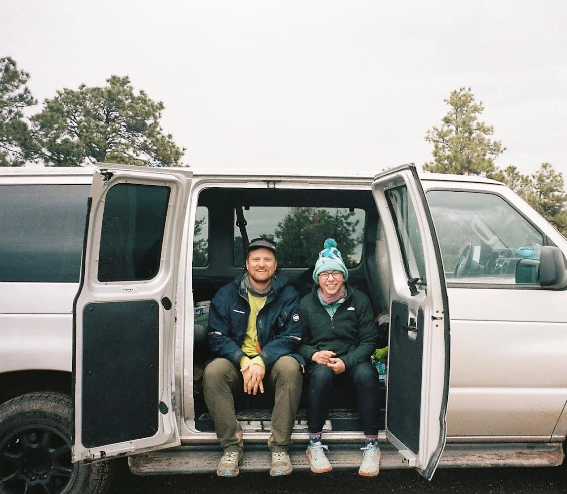Two people sitting inside a silver van with open sliding doors, outdoors with trees in the background, smiling and dressed in outdoor clothing.