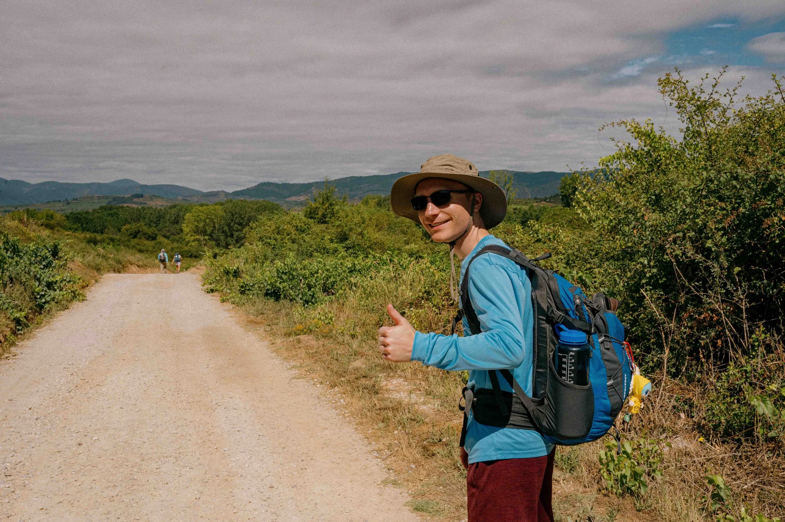 A young man hiking on a dirt trail in a scenic landscape, giving a thumbs up and smiling, wearing sunglasses, a wide-brimmed hat, a blue long-sleeve shirt, red pants, and carrying a backpack with water bottles, with other hikers in the distance and mountains in the background.