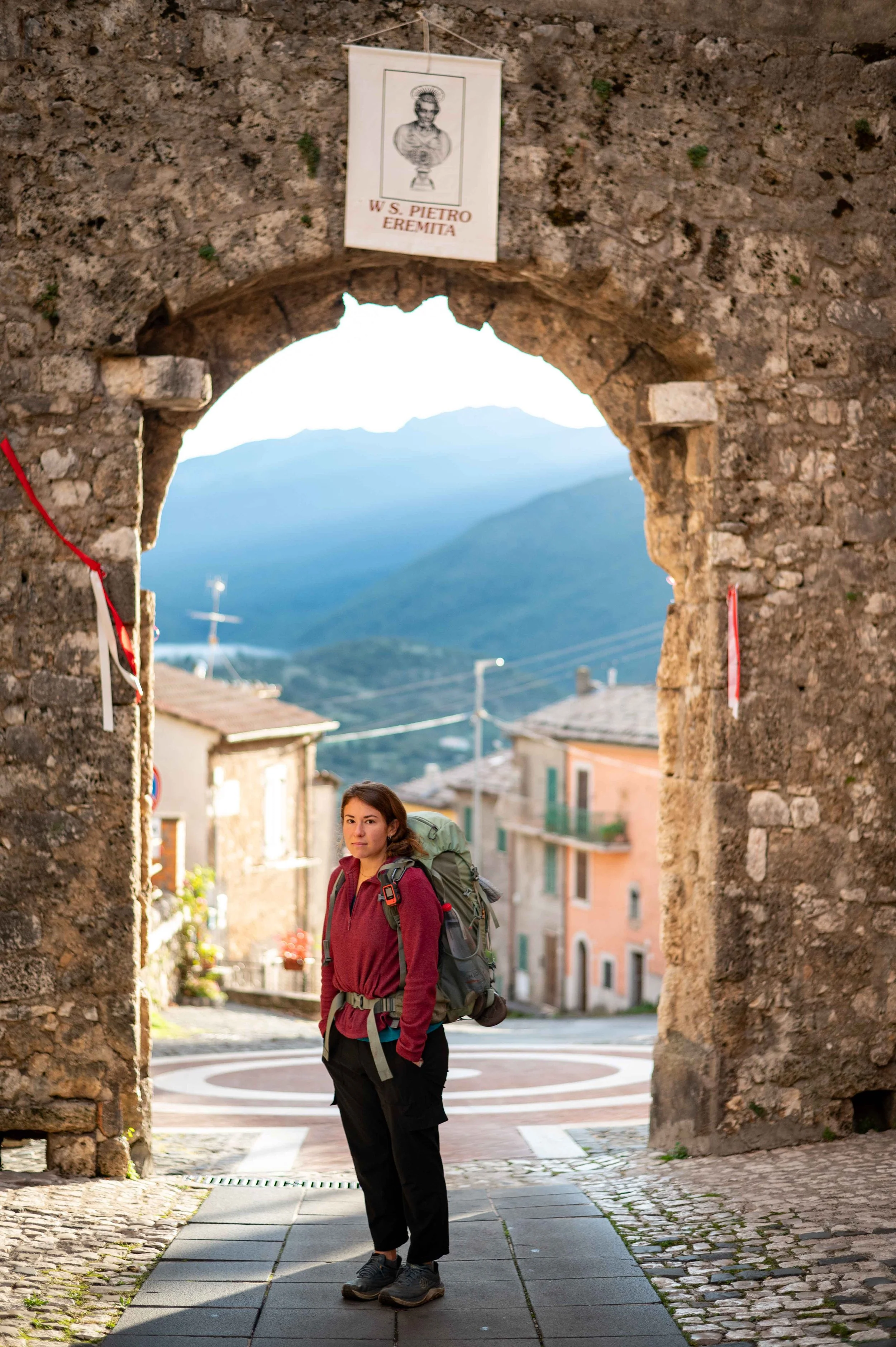 A young woman with a backpack standing in an old stone archway in a small Italian village with mountains in the background.