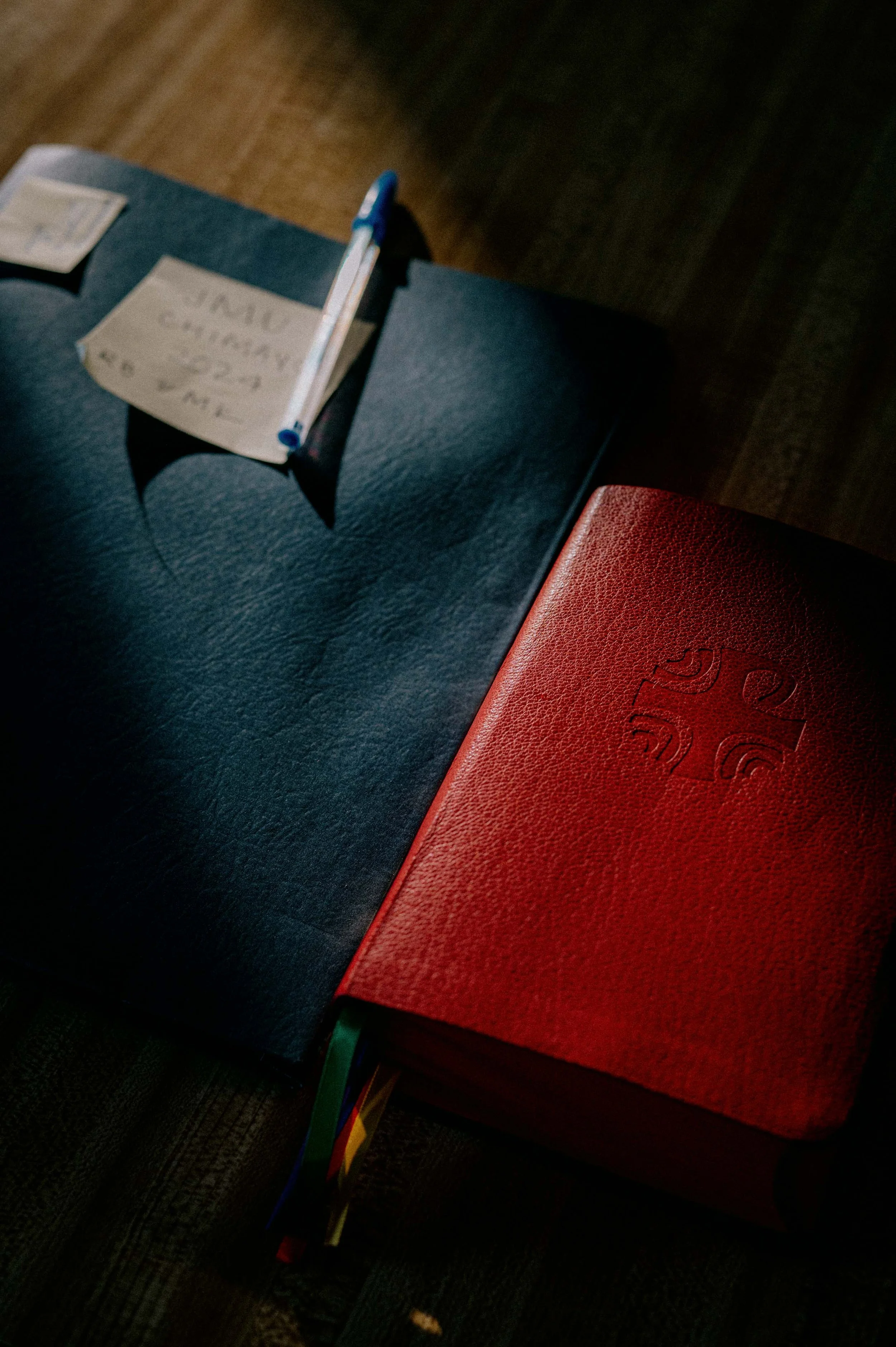 A red leather Bible with an embossed cross on the cover, placed on a black book, with a white note and a blue pen on top of the black book, sitting on a dark wooden surface.