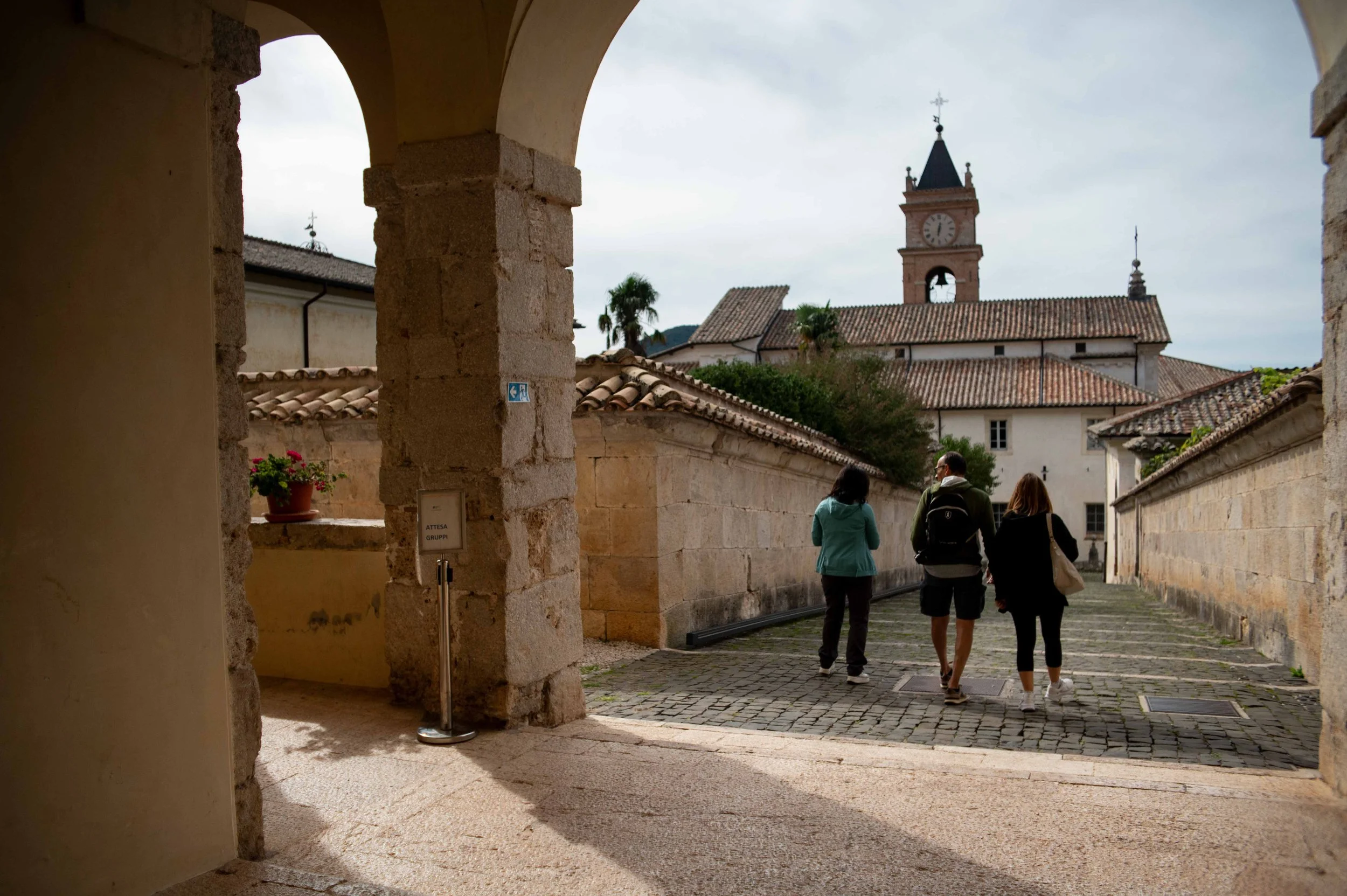 Three people walking down a cobblestone street in a historic town with old stone buildings and a bell tower in the background.