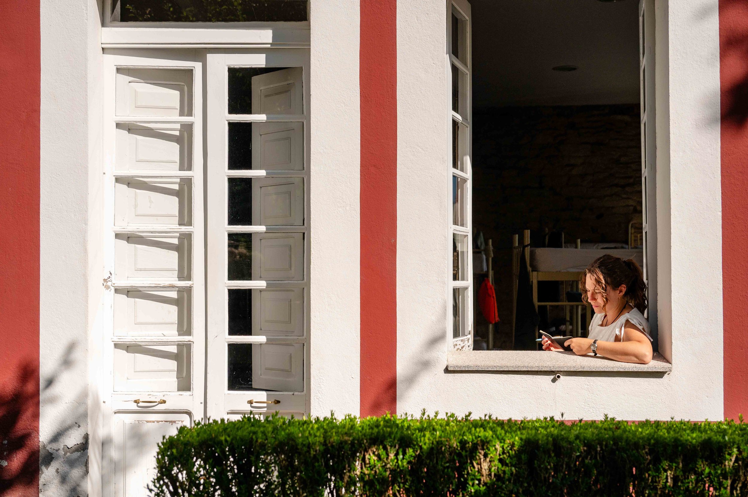 A woman sitting at an open window, looking at her phone, with greenery in front and part of a pink and white building.