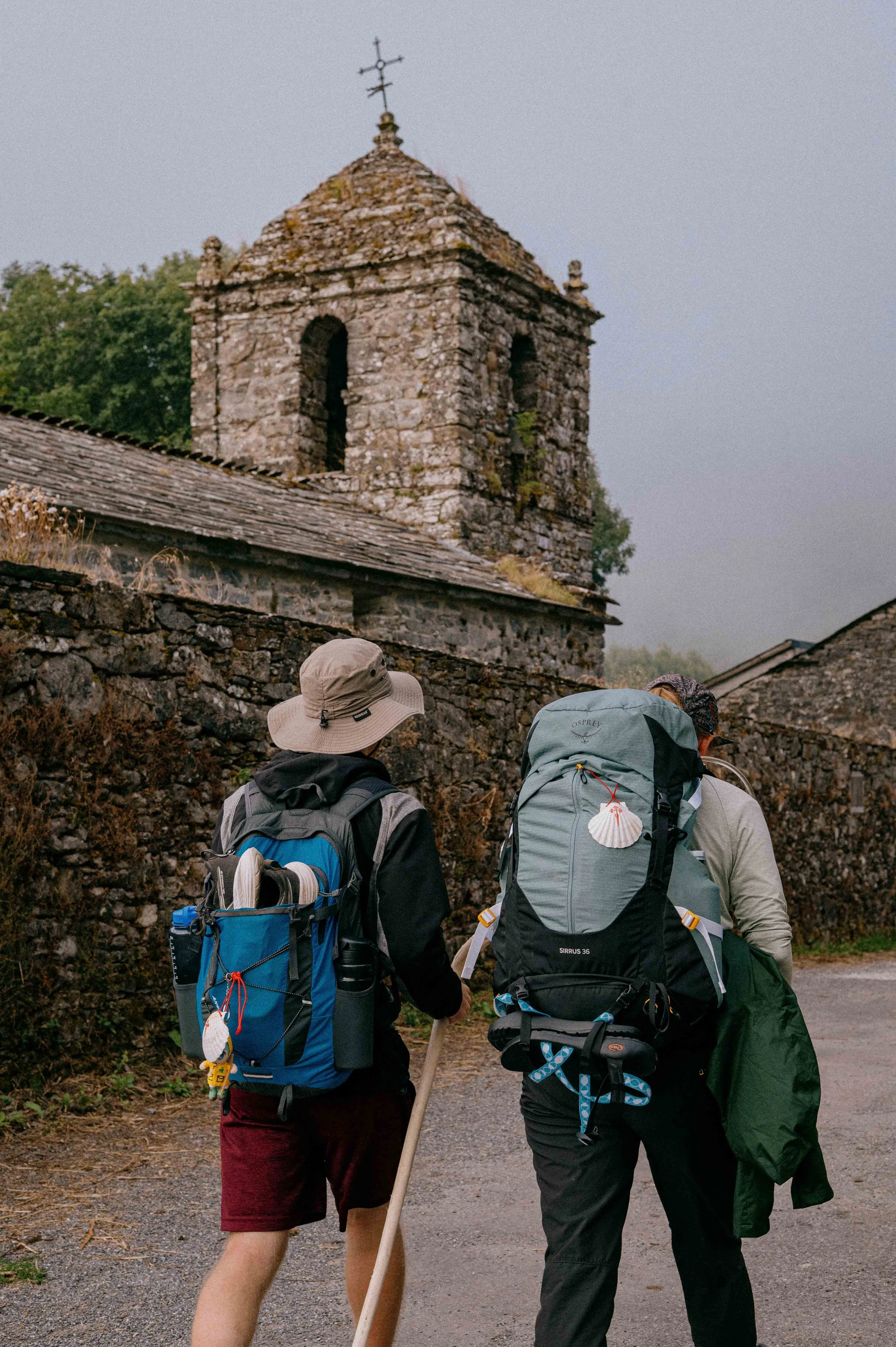 Two hikers with backpacks walking along a rural stone path with an old stone church in the background under a cloudy sky.