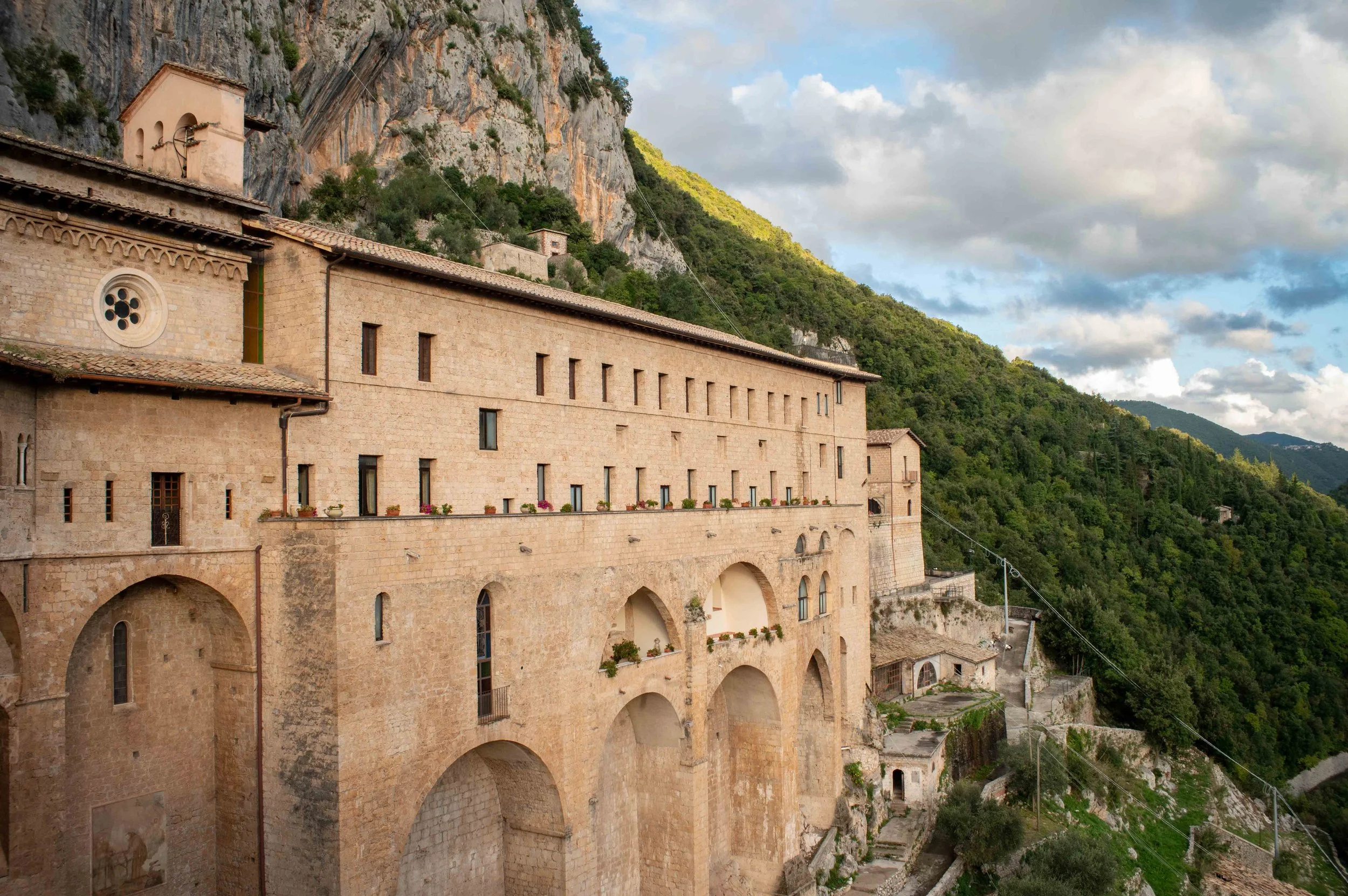 Stone building built into the side of a mountain with arches and small windows, with lush green hills and a partly cloudy sky in the background.