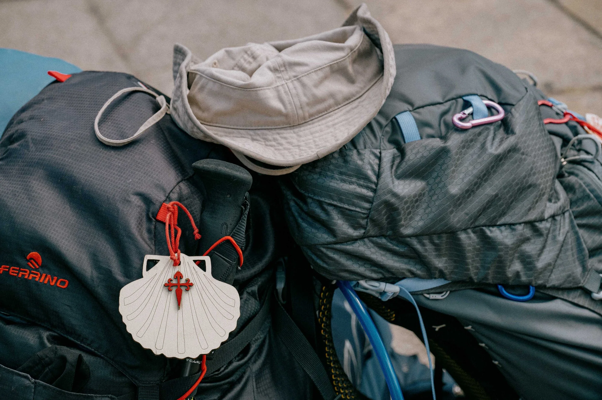 Close-up of two hiking backpacks with gear, including a beige hat placed on top, and a hanging tag shaped like a shell with a red cross symbol.