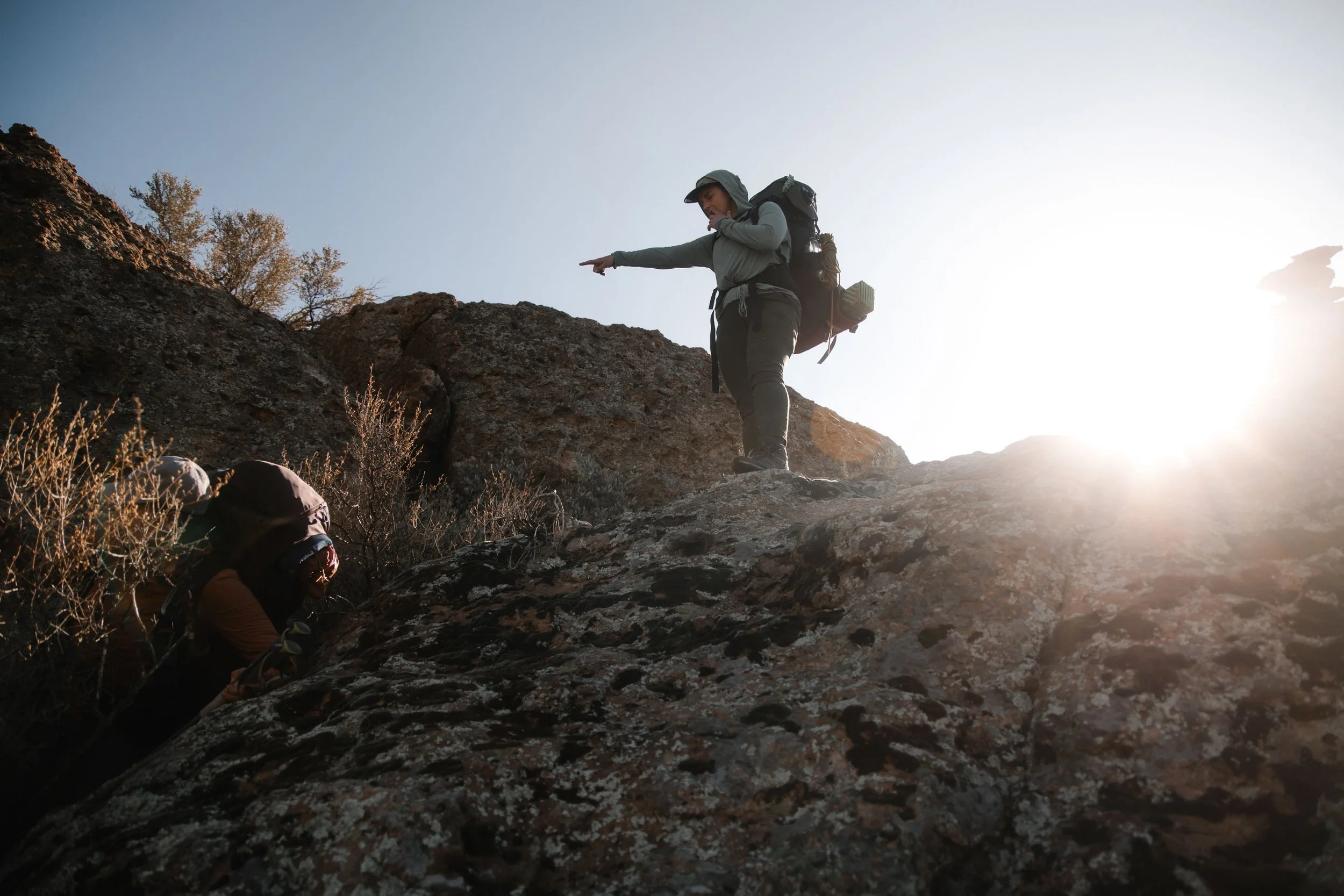 Two hikers, one climbing up a rocky incline and the other standing on a higher ledge, outdoors during a sunny day