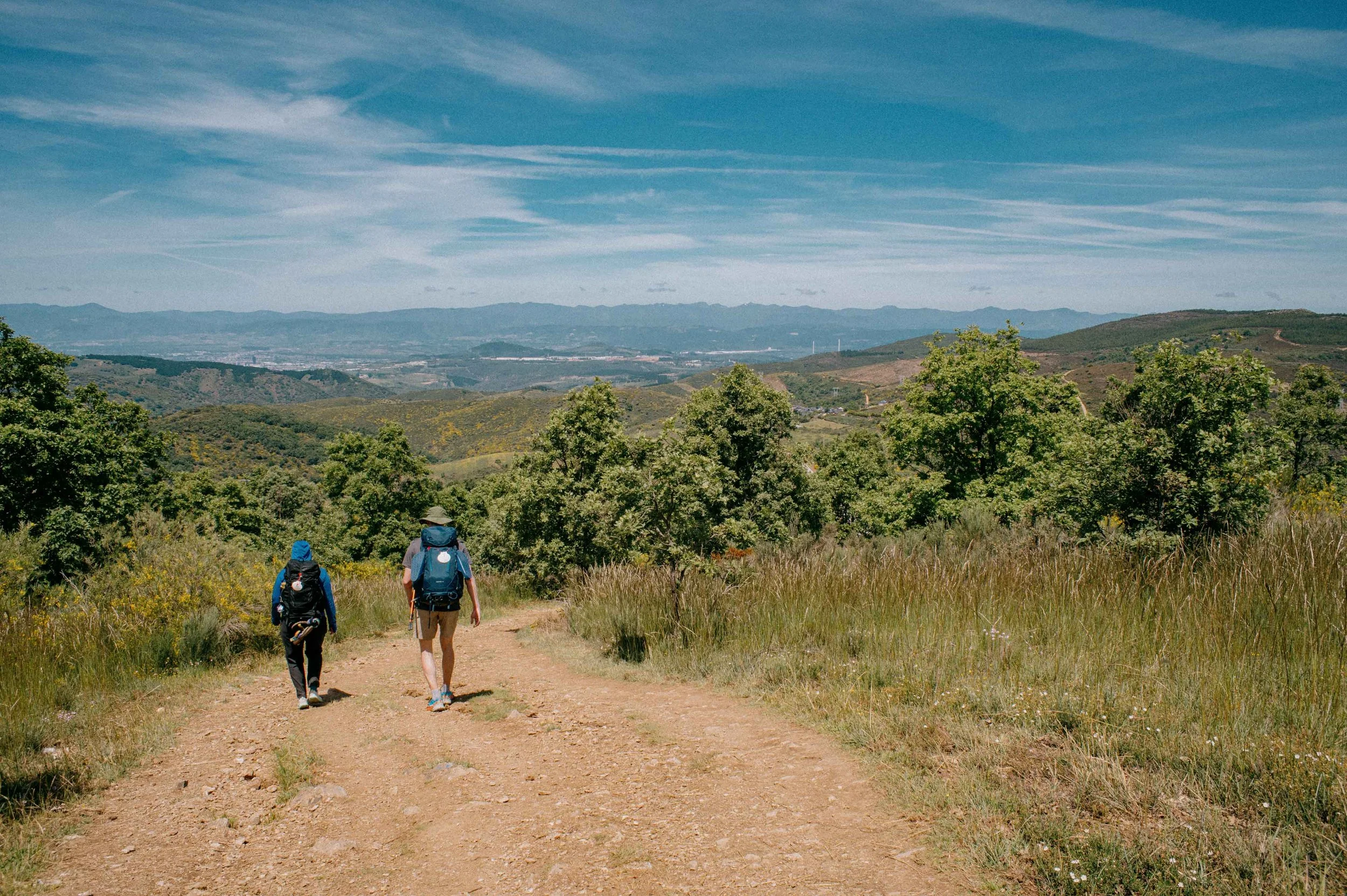 Two hikers walking on a dirt trail through a green, hilly landscape under a partly cloudy blue sky.