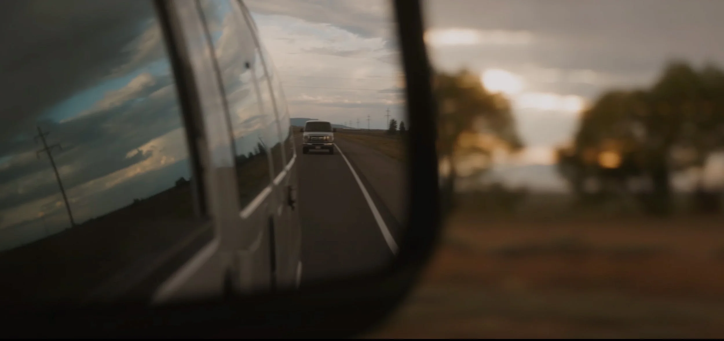 A view through a vehicle's side mirror showing a road with a car approaching, with trees and power lines in the background, during sunset or dusk.