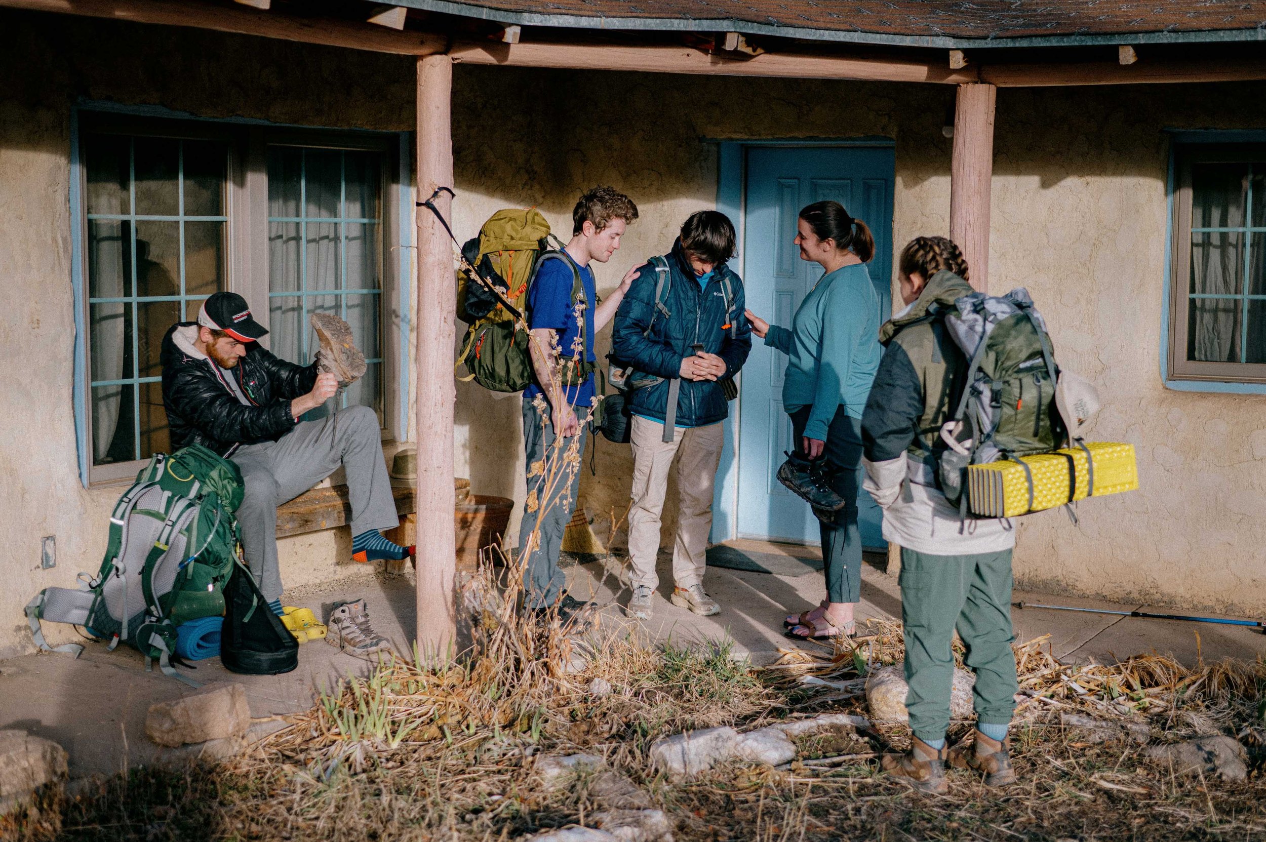 Five hikers, three men and two women, standing outside a house with blue door and window frames, preparing for a hike. One man is sitting on a step adjusting his shoes, while the others are standing and talking, with backpacks and gear.