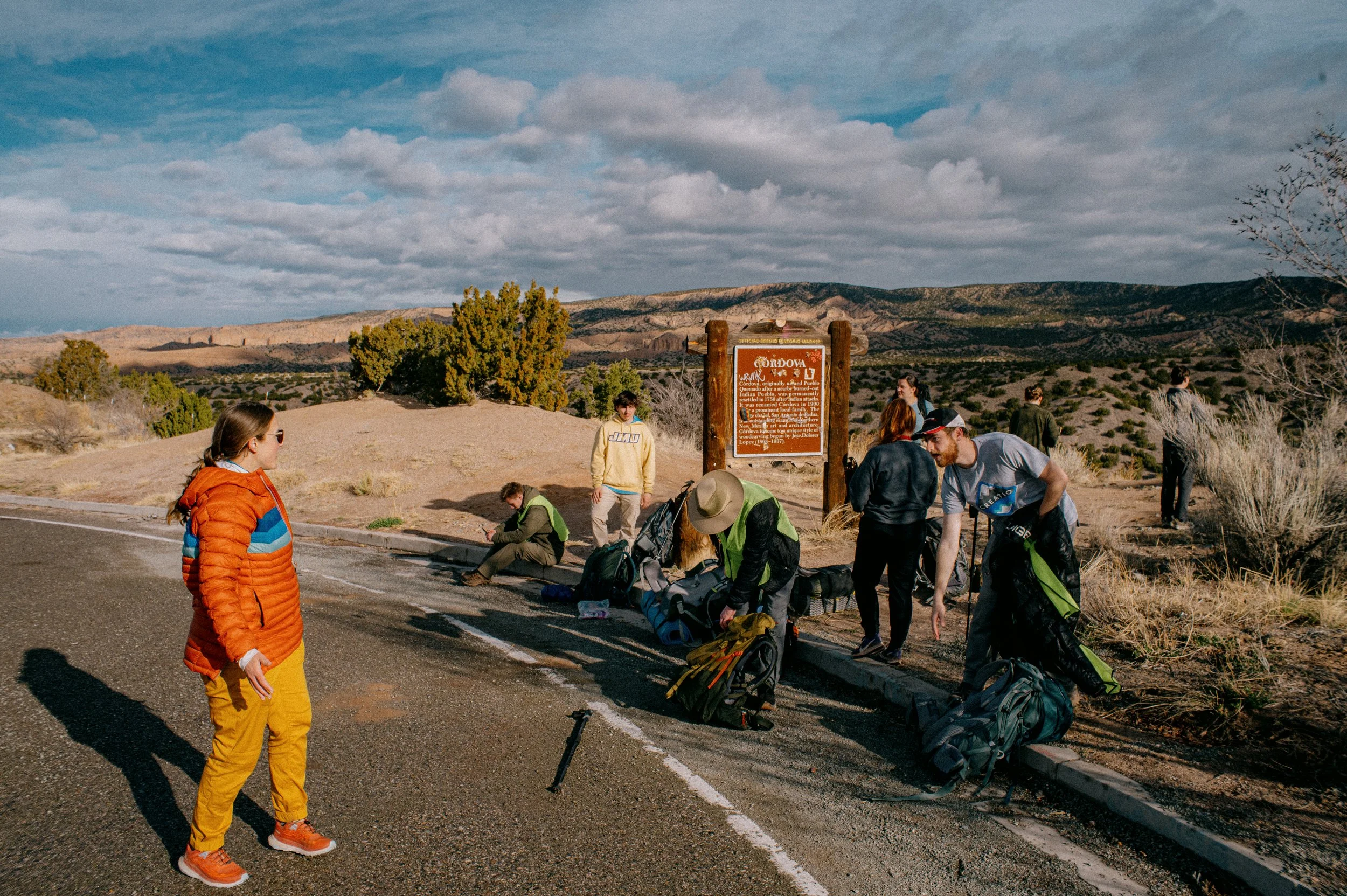 People gathering with backpacks and gear near a sign in a desert landscape, preparing for an outdoor activity, with mountains and a cloudy sky in the background.