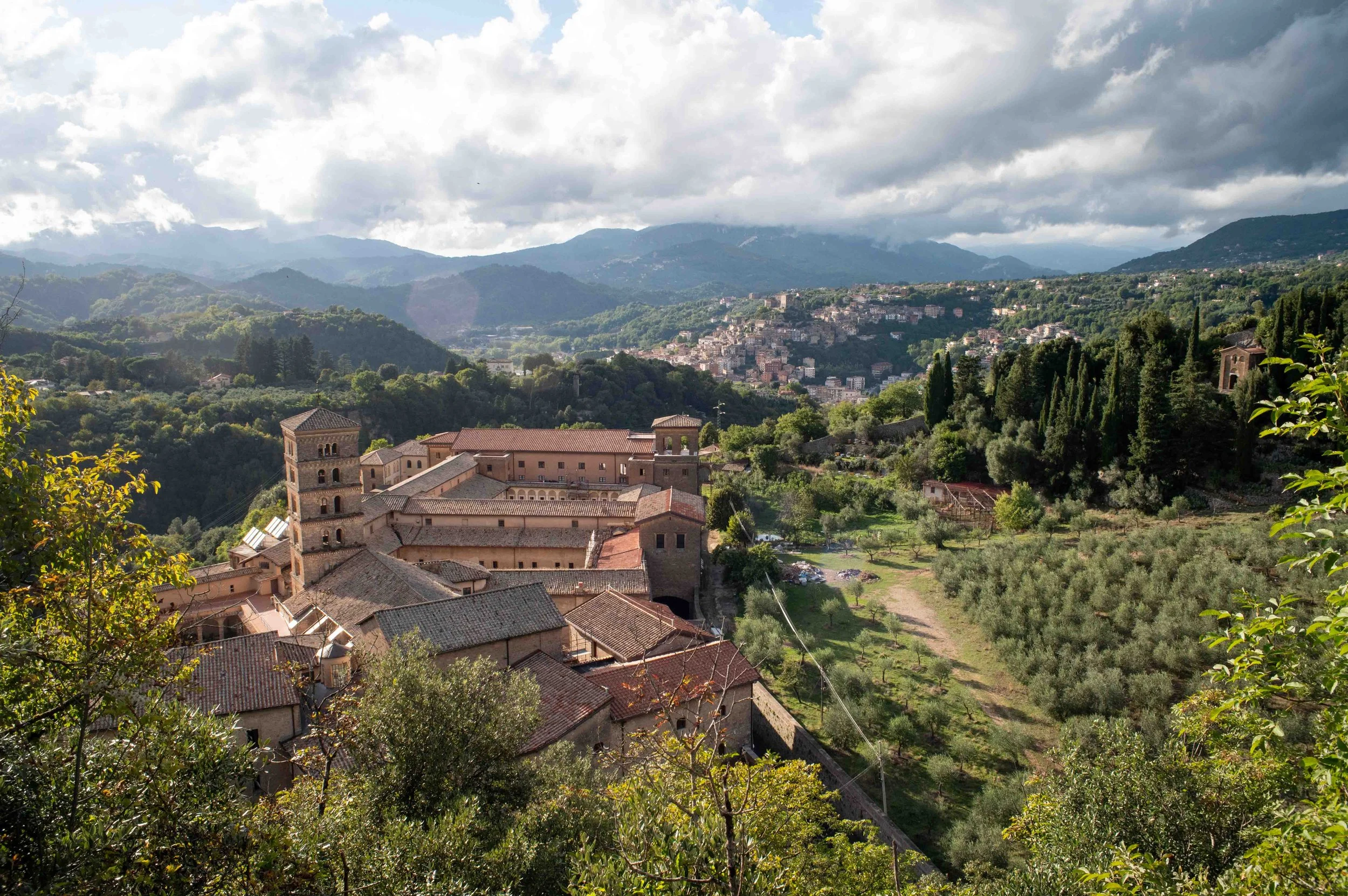 Scenic view of a hillside town with historic brick buildings, green trees, and mountains in the background under partly cloudy sky.