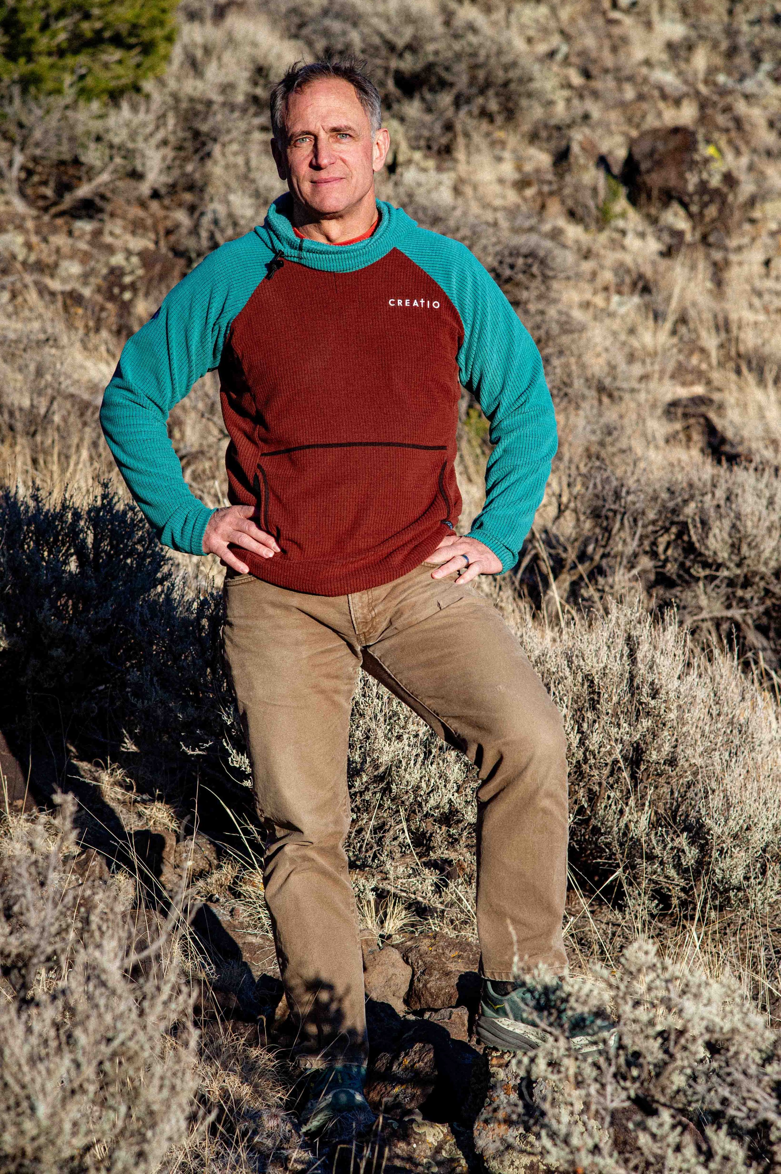 A man standing outdoors on rocky terrain with dry bushes and mountains in the background, wearing a color-blocked turquoise and red Creatomic hoodie, beige pants, and hiking shoes.