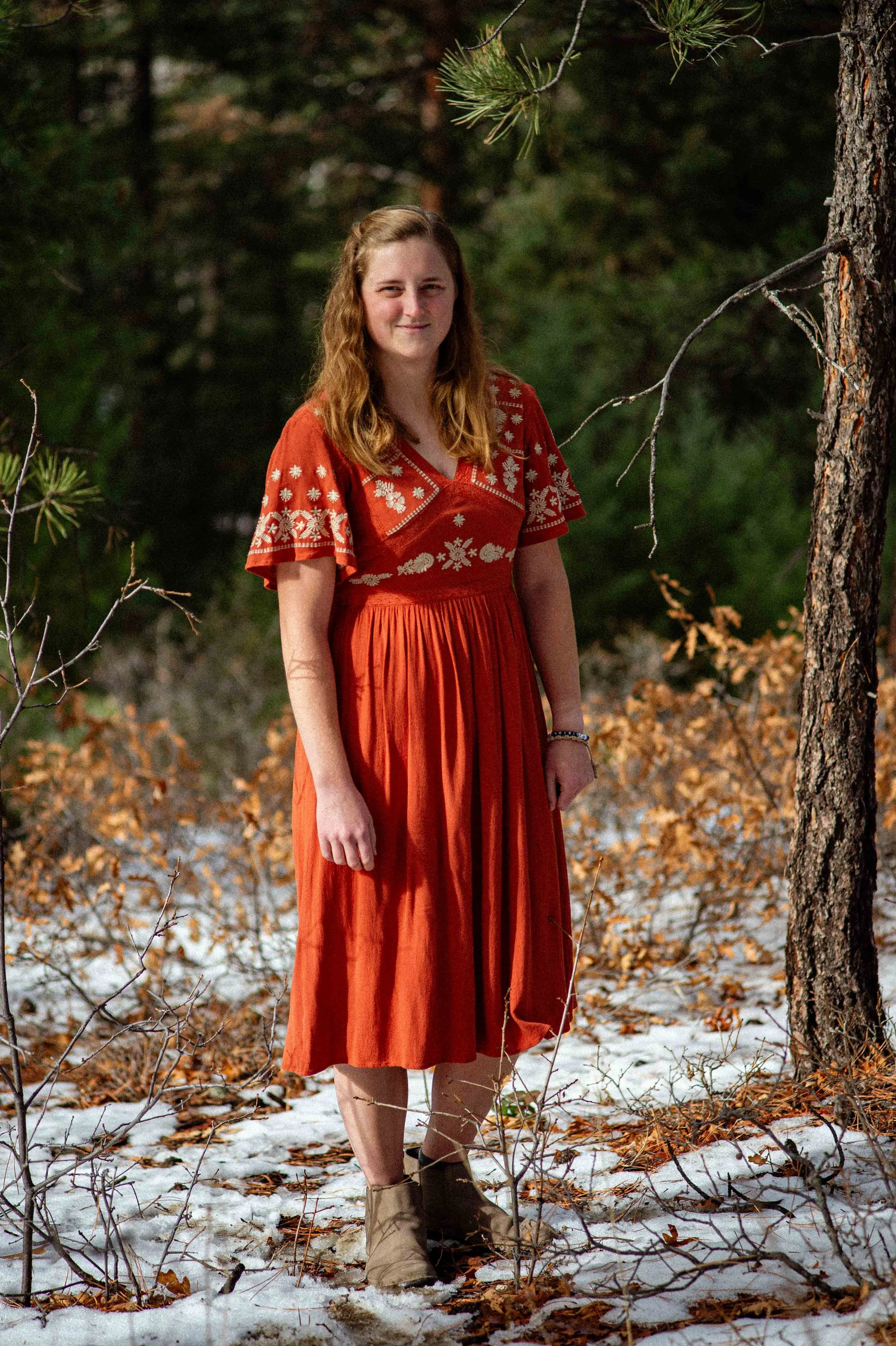 A young woman with long, wavy red hair stands outdoors in a forested area with snow on the ground and fallen leaves. She is wearing a rust-colored dress with embroidered white patterns on the sleeves and chest, and tan boots. She is smiling softly and looking at the camera.