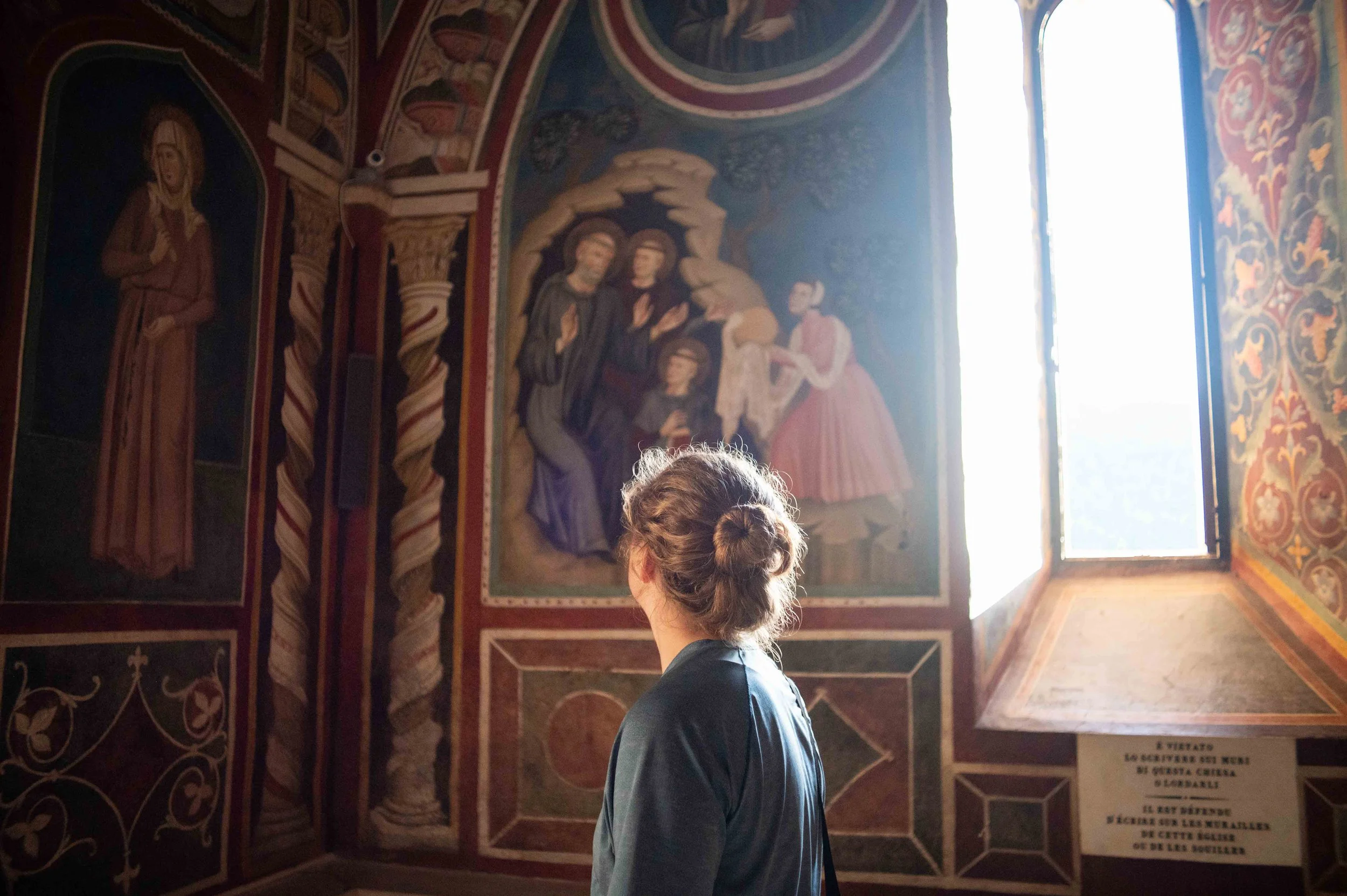A woman with curly hair tied in a bun standing in front of religious mural paintings inside a church, with sunlight coming through a window on the right side.