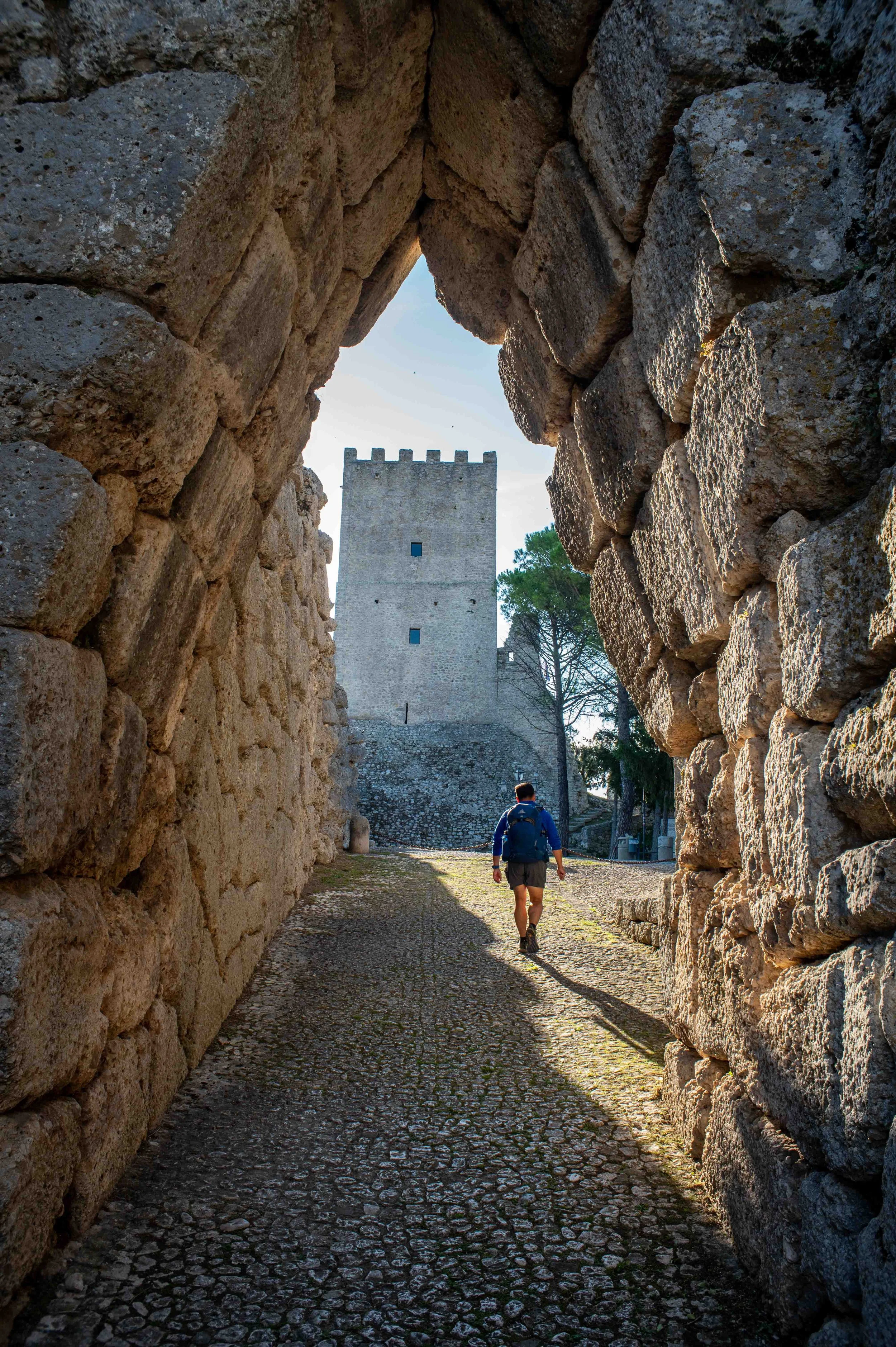 A person walking through a stone archway of an ancient castle or fortress with a tall rectangular tower visible in the background, and sunny weather.