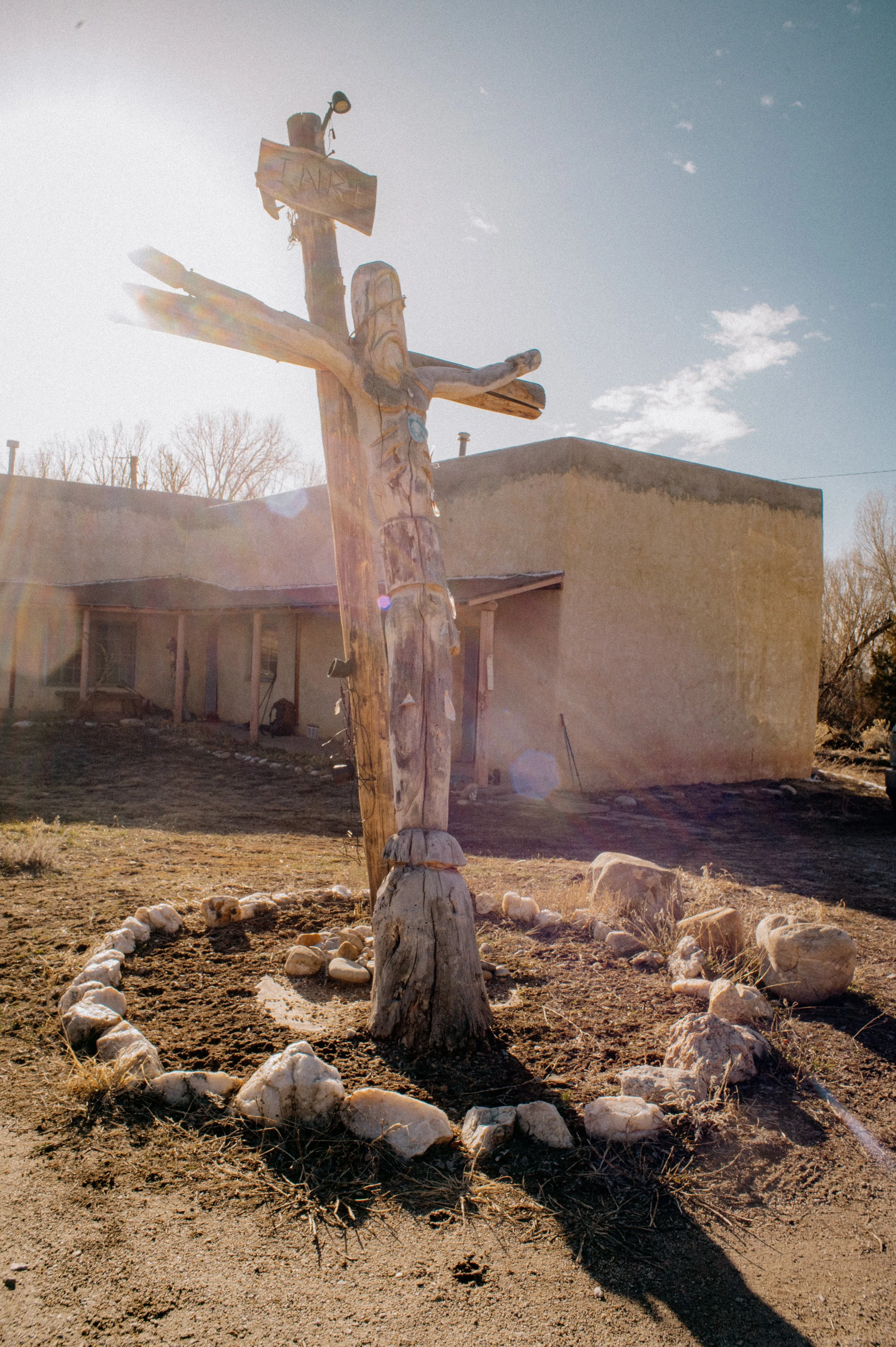 A wooden cross with a figure of Jesus Christ carved from wood, surrounded by rocks and situated outside a stucco building, with the sun shining behind it.