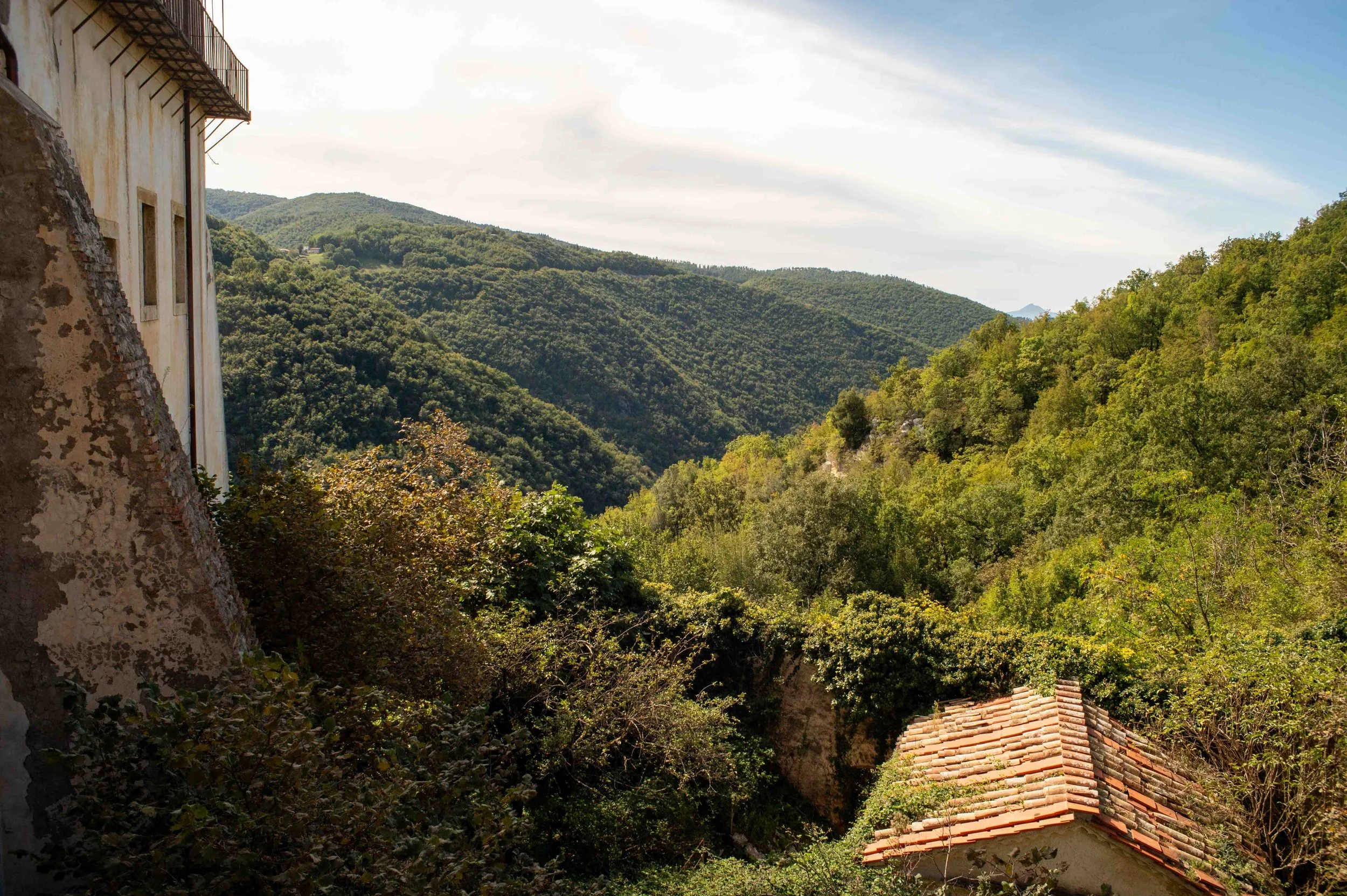 A scenic view of lush green mountains with a small building with a red-tiled roof in the foreground, partially covered by trees and bushes, under a partly cloudy sky.