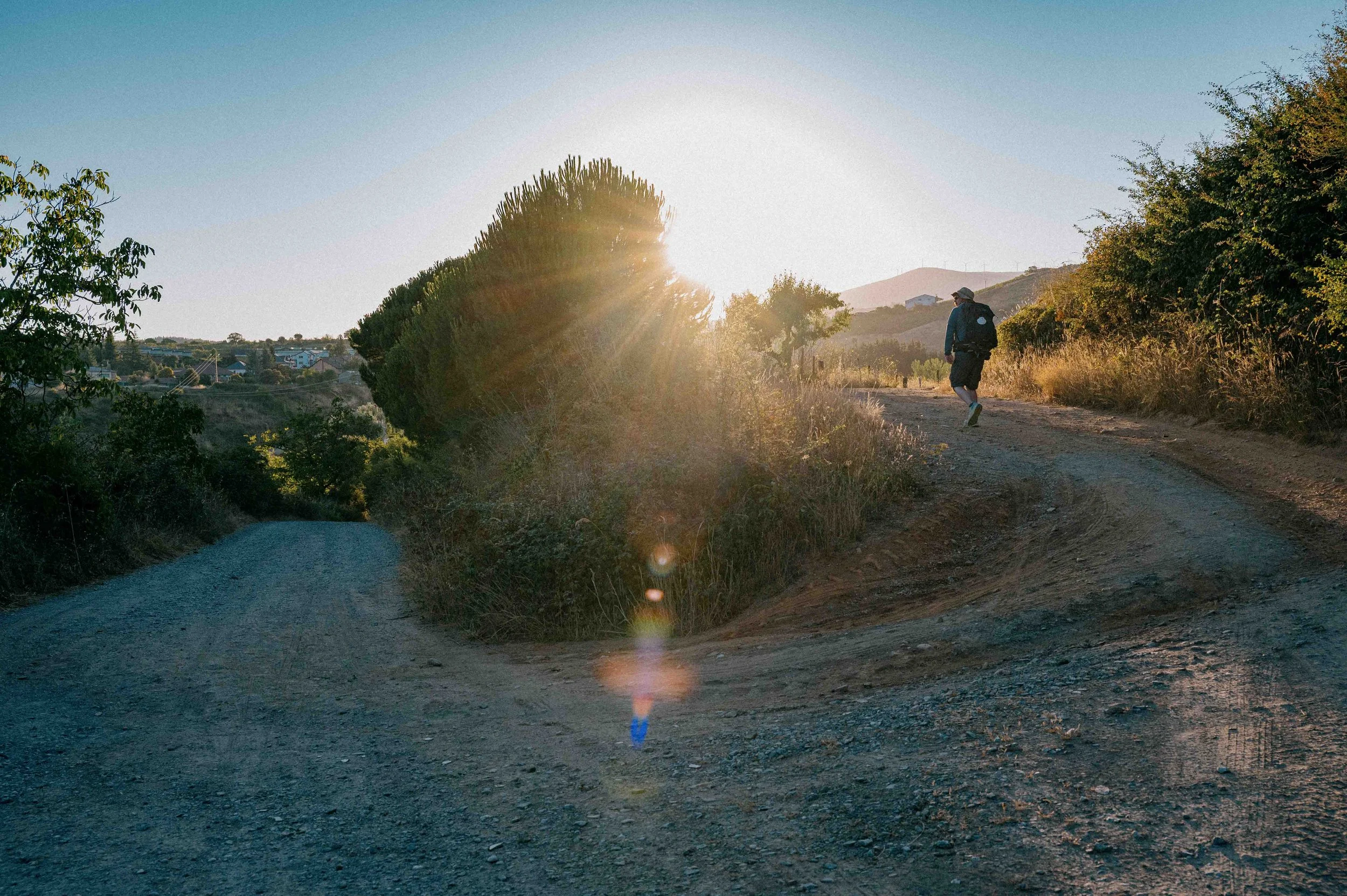 A person hiking on a dirt trail in a hilly area during sunset, with trees and shrubs on either side.