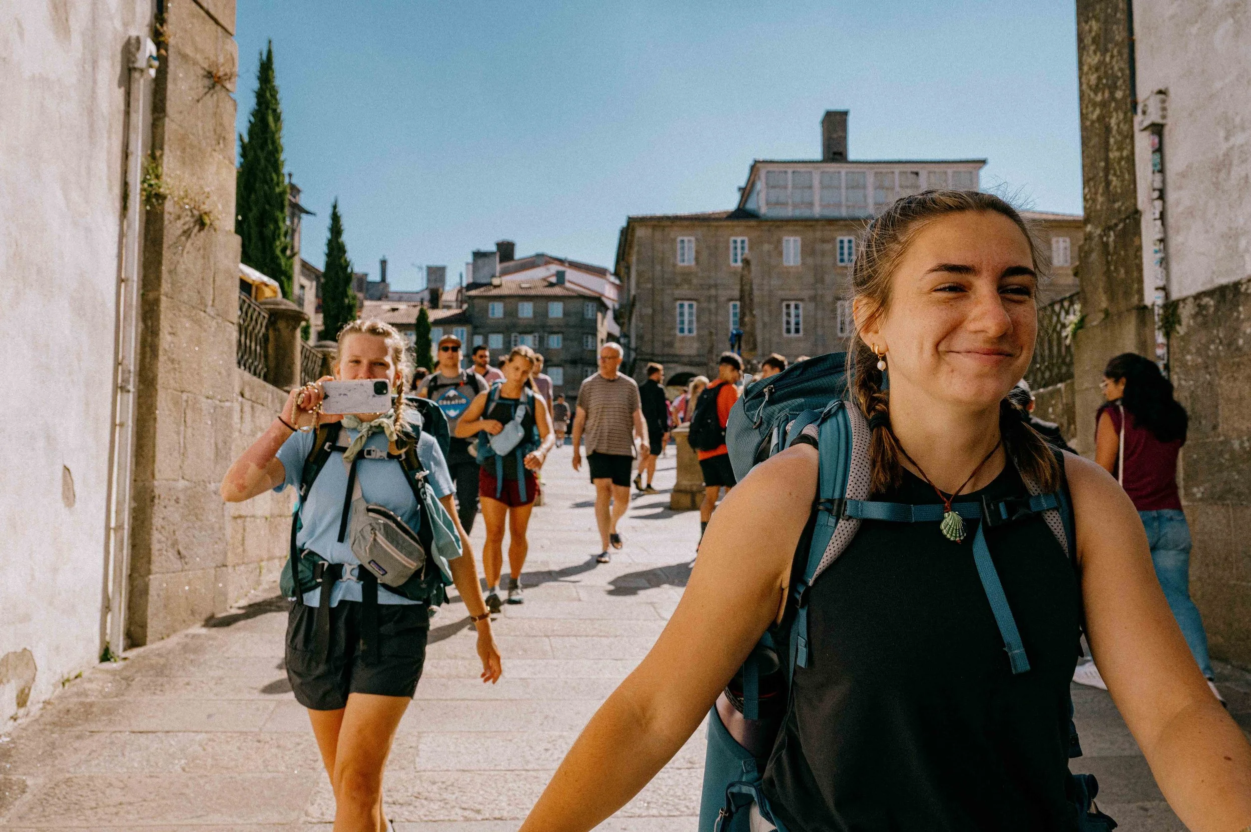 Group of young backpackers walking up a city street with historic stone buildings and clear blue sky.