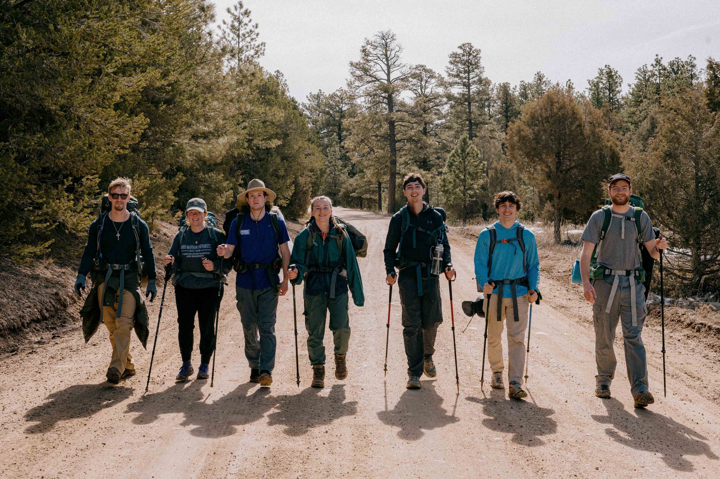 Group of seven hikers walking on a dirt trail in a forested area, carrying backpacks and hiking poles on a sunny day.