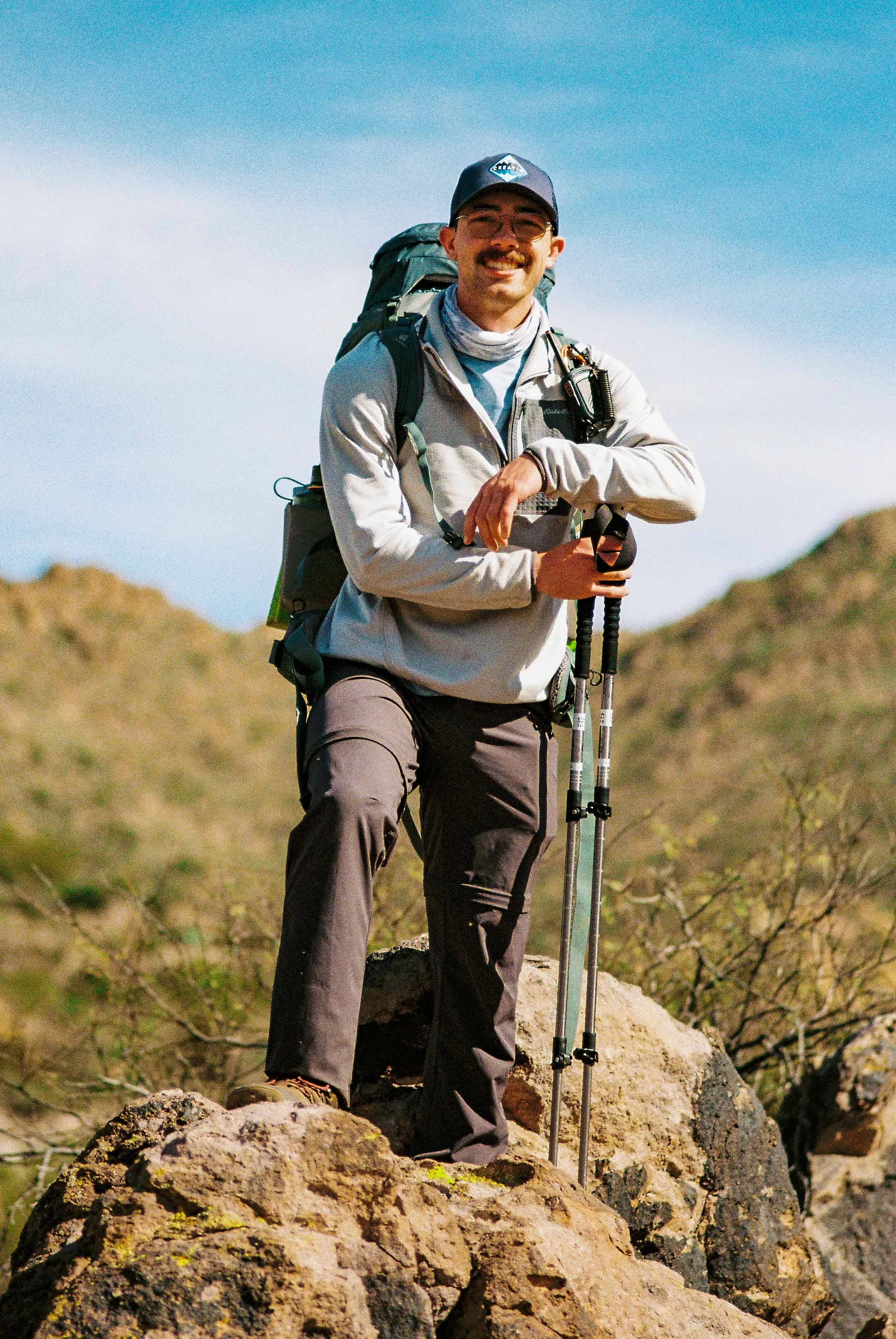 A man hiking outdoors, wearing a gray jacket, dark pants, sunglasses, and a baseball cap, holding trekking poles and smiling. He has a backpack and is standing on a rocky terrain with hills in the background under a blue sky.