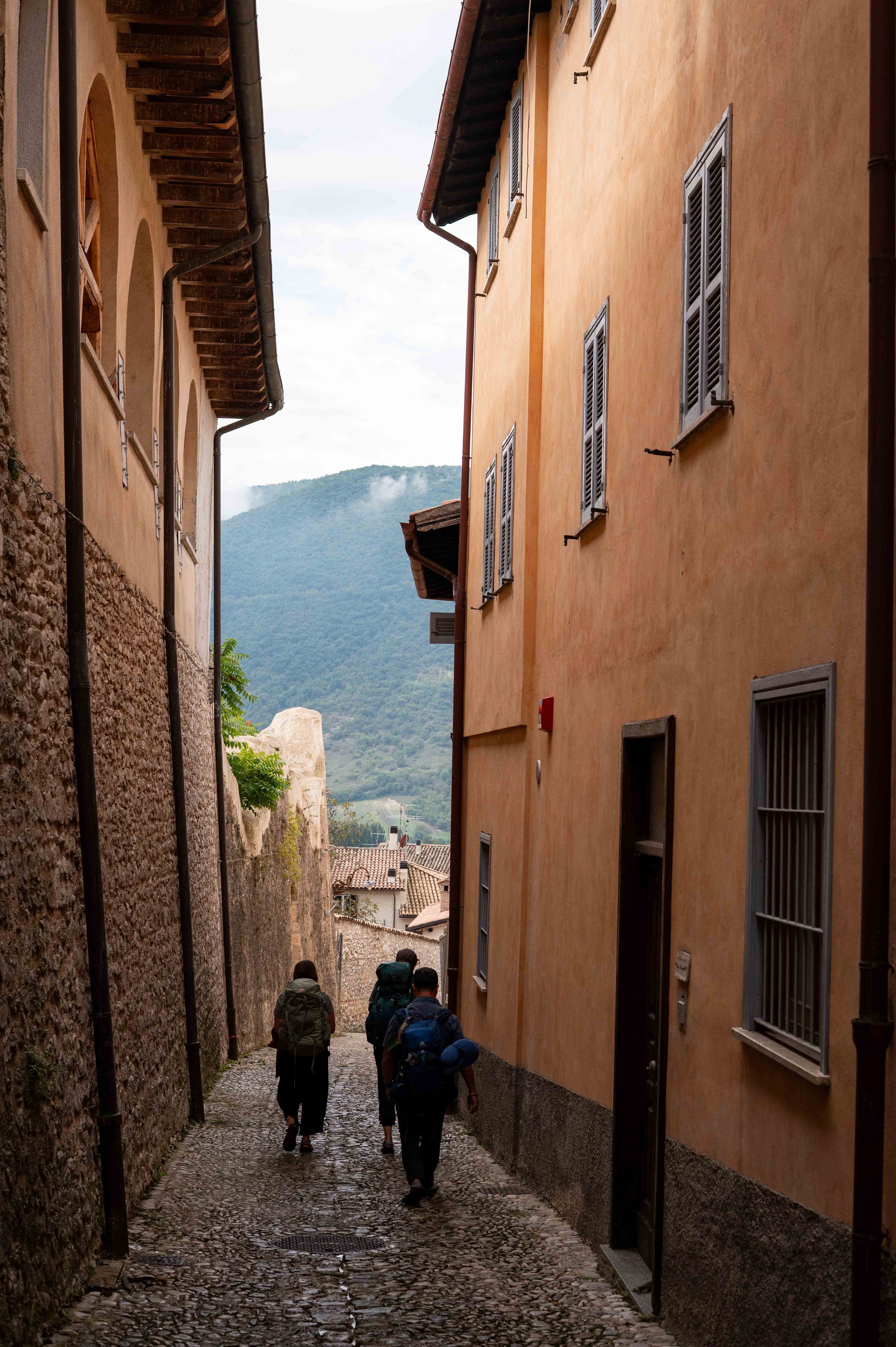 A narrow cobblestone alleyway flanked by tall, colorful buildings with shuttered windows, with mountains in the background and four hikers walking away from the camera.