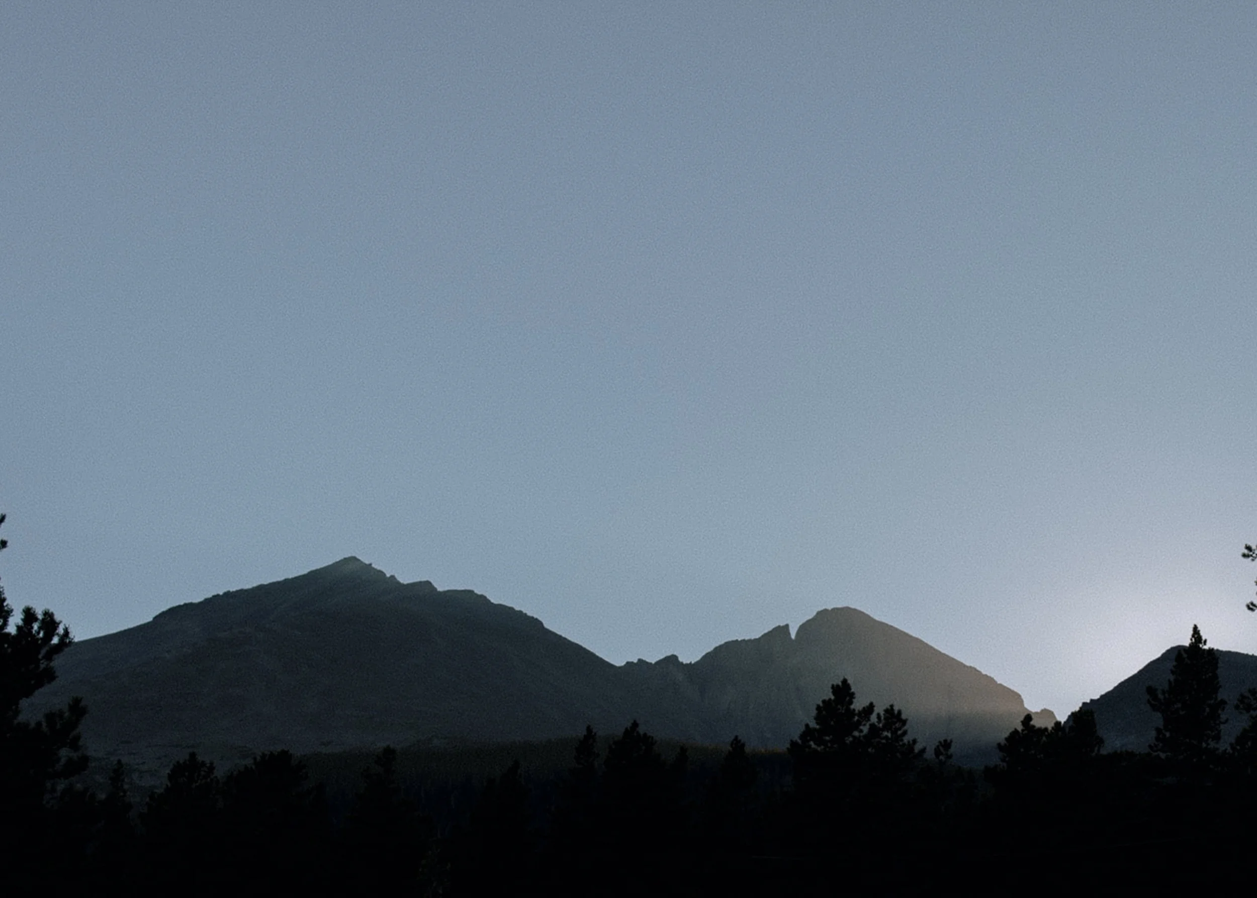 Silhouette of a mountain range at dusk with a clear sky above and tree silhouettes in the foreground.