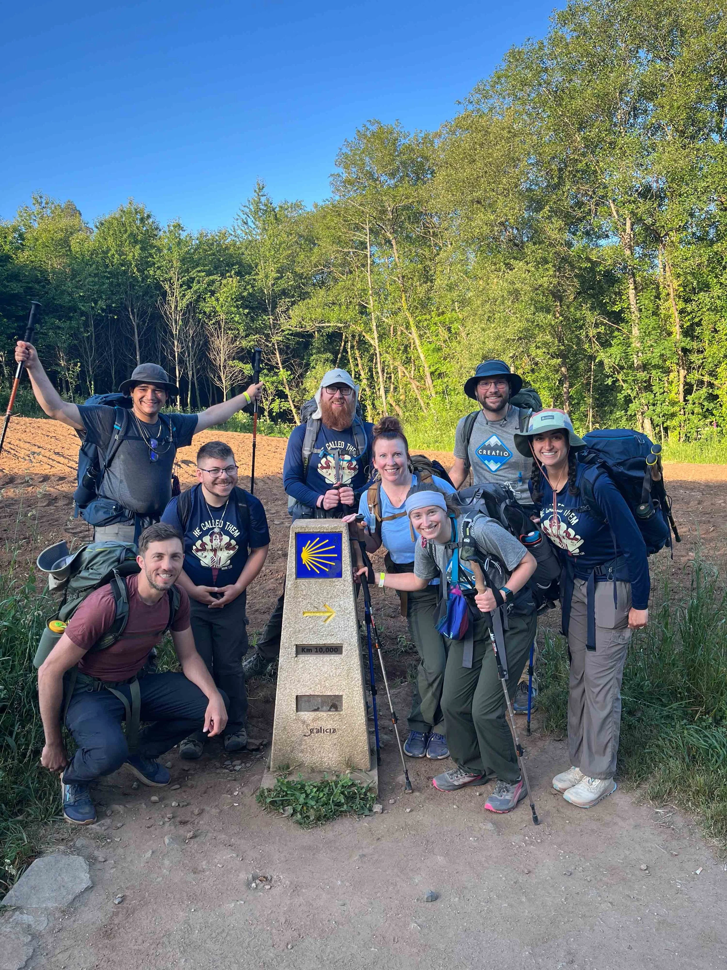 Group of hiking volunteers smiling near a milestone marker with a yellow shell symbol, on a trail surrounded by trees and greenery under a clear blue sky.