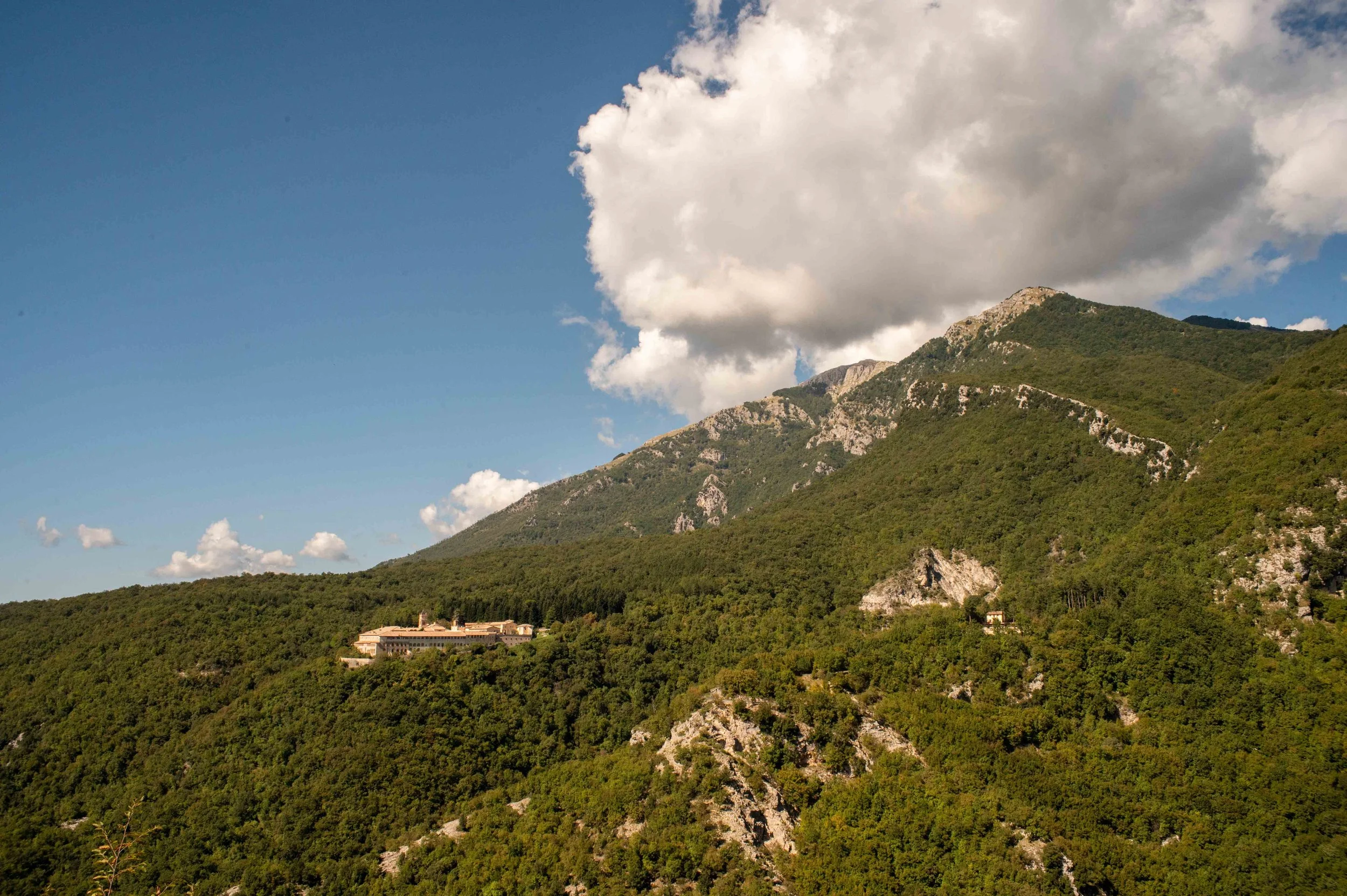 Mountain landscape with lush green trees, rocky slopes, a building near the base, and a partly cloudy sky.