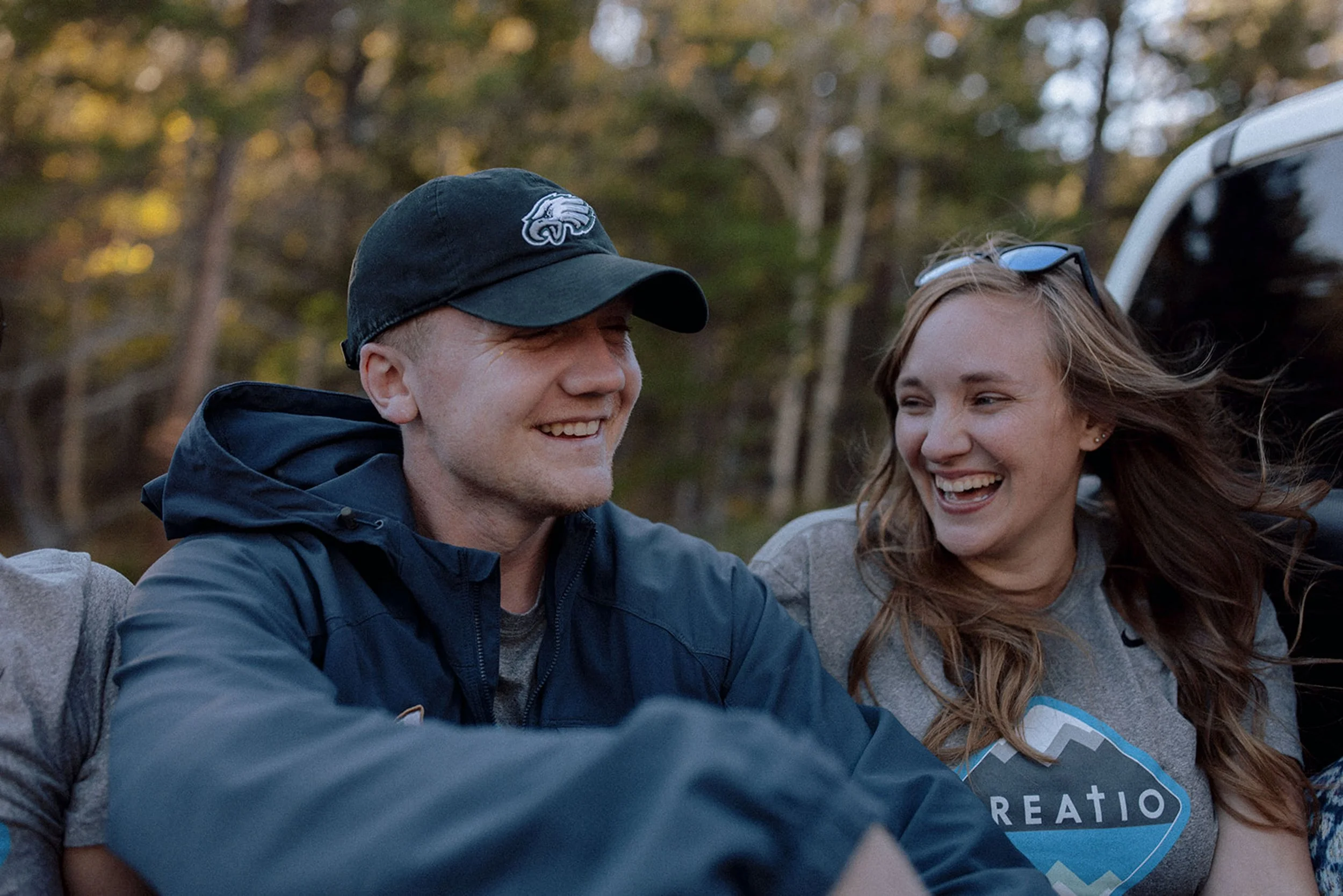 A young man and woman smiling and sharing a happy moment outdoors in a wooded area, with trees and a vehicle in the background, during daylight.