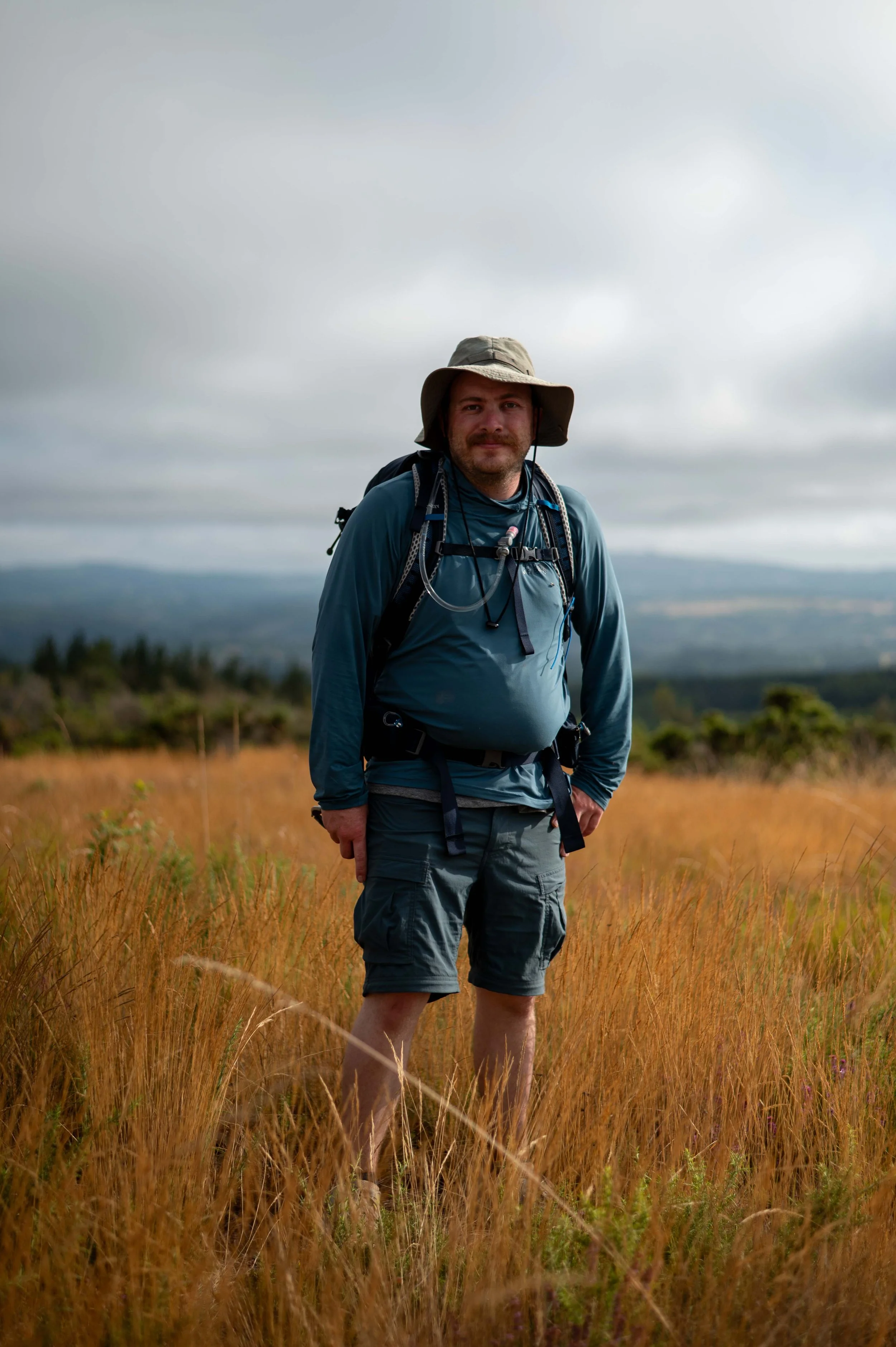 A man standing in a grassy field with a cloudy sky, wearing outdoor hiking gear including a hat, a backpack, and shorts.