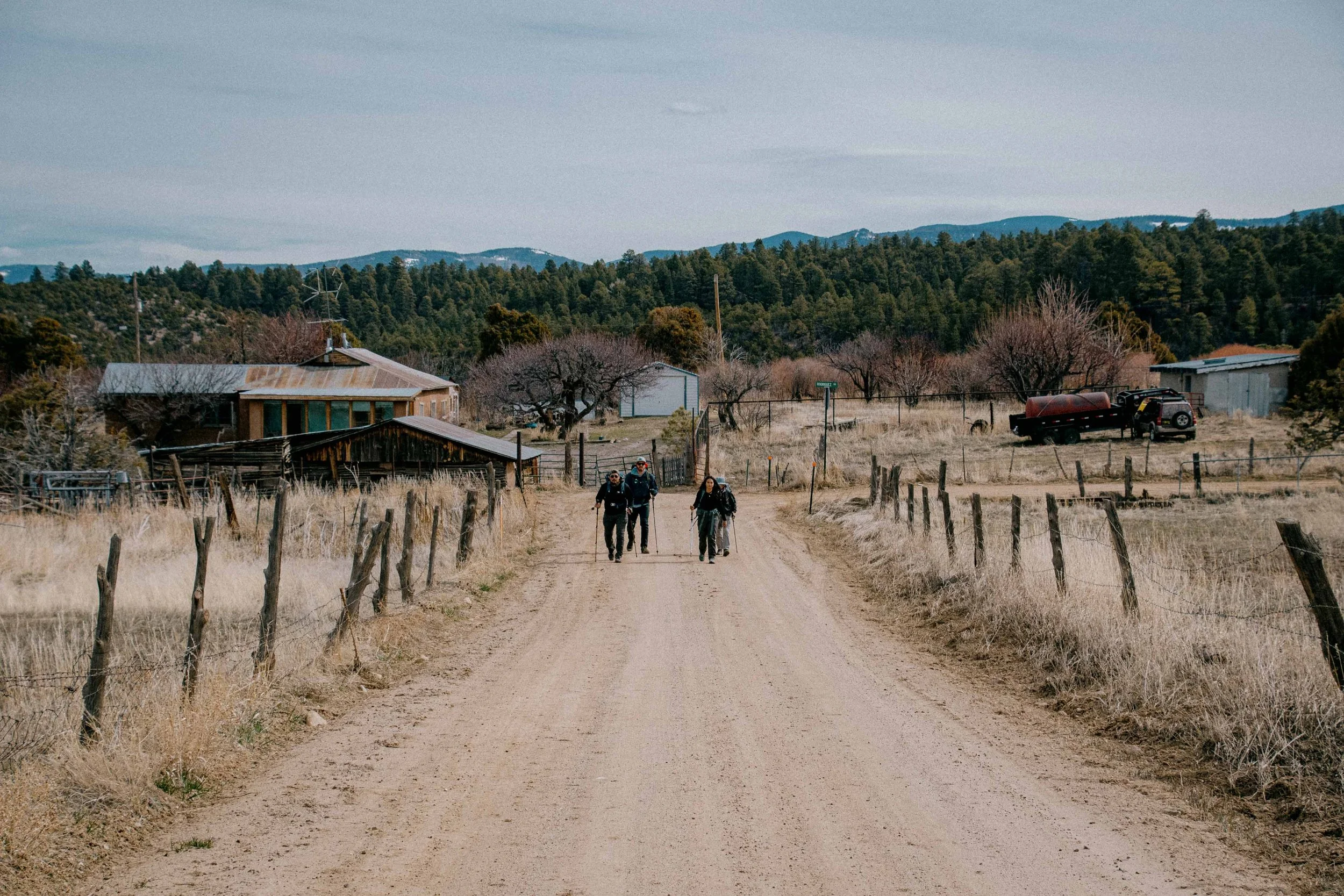 Three hikers walking down a dirt rural road with fences on both sides, scattered houses, leafless trees, and distant mountains in the background.