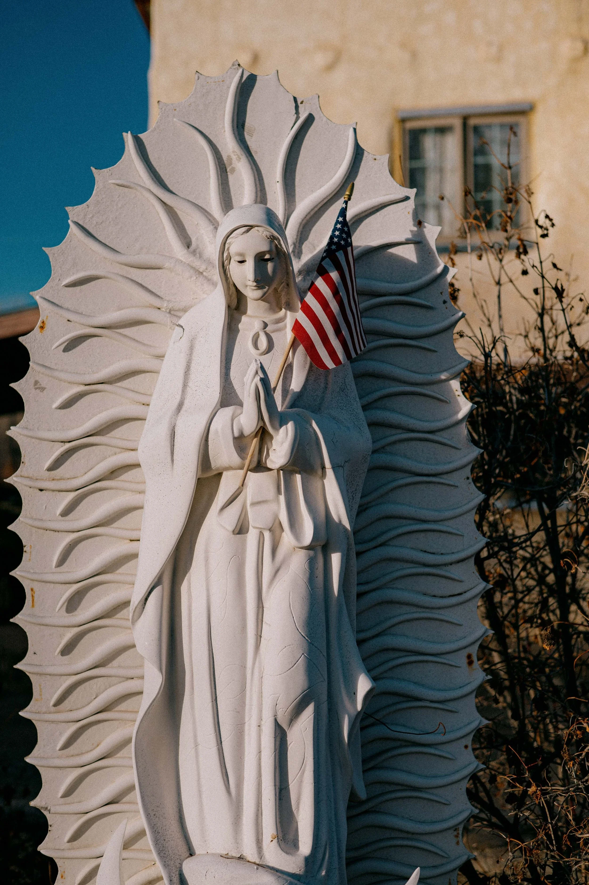 Statue of the Virgin Mary holding an American flag, with a decorative white backdrop resembling a flower or angel wings. The background includes a building with a window and some dried plants.