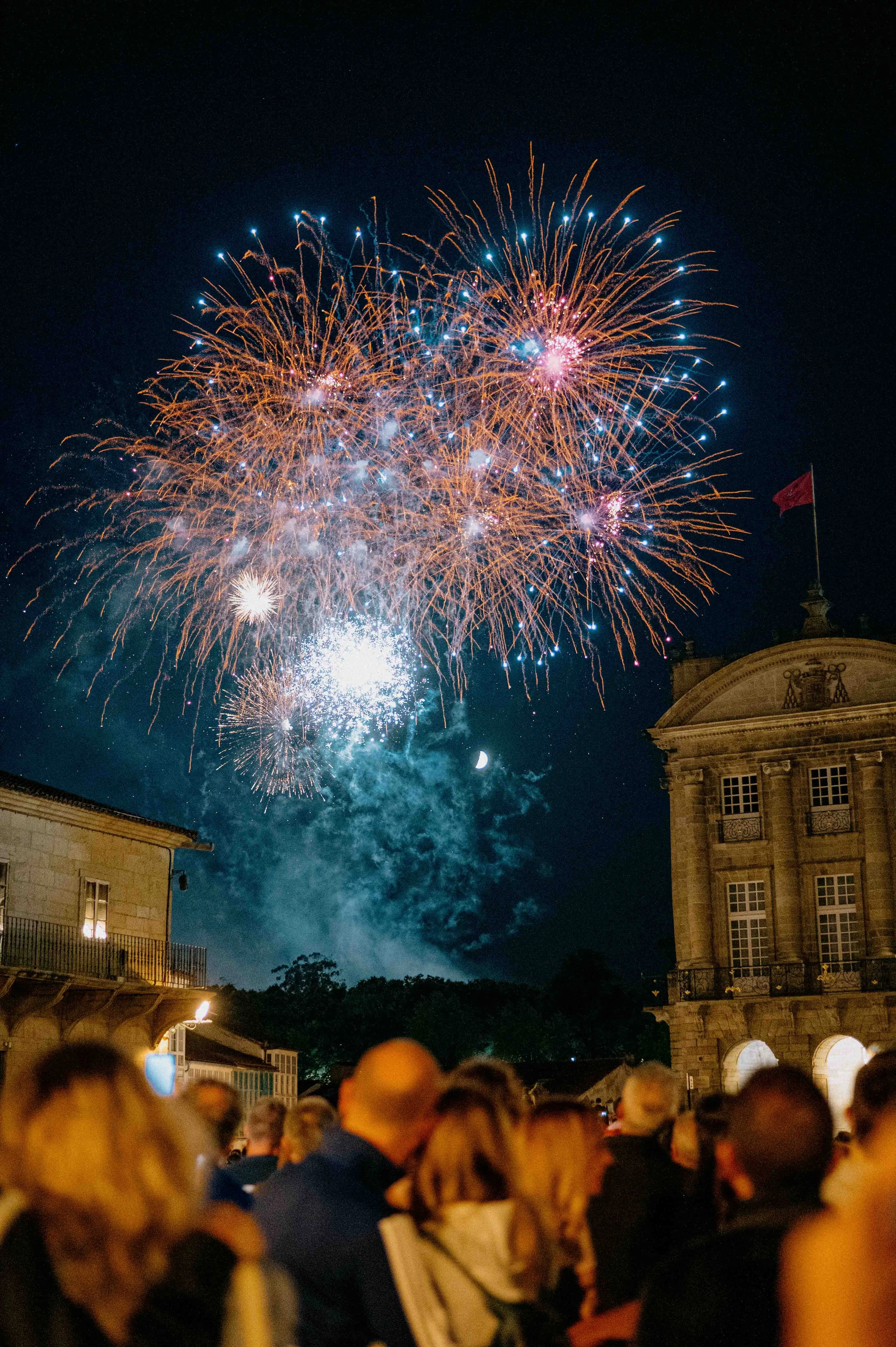 People watching fireworks display at night with a historic building in the background and a crescent moon in the sky.