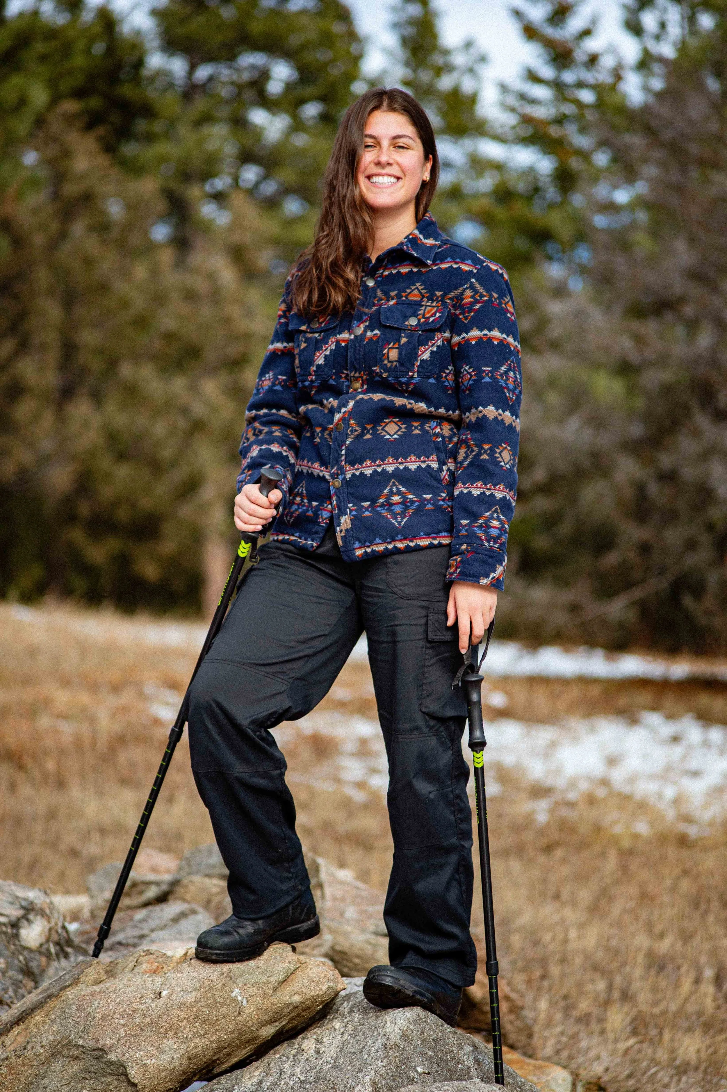 Young woman with long brown hair wearing a patterned jacket, black cargo pants, and hiking boots, standing on rocks outdoors holding trekking poles and smiling.