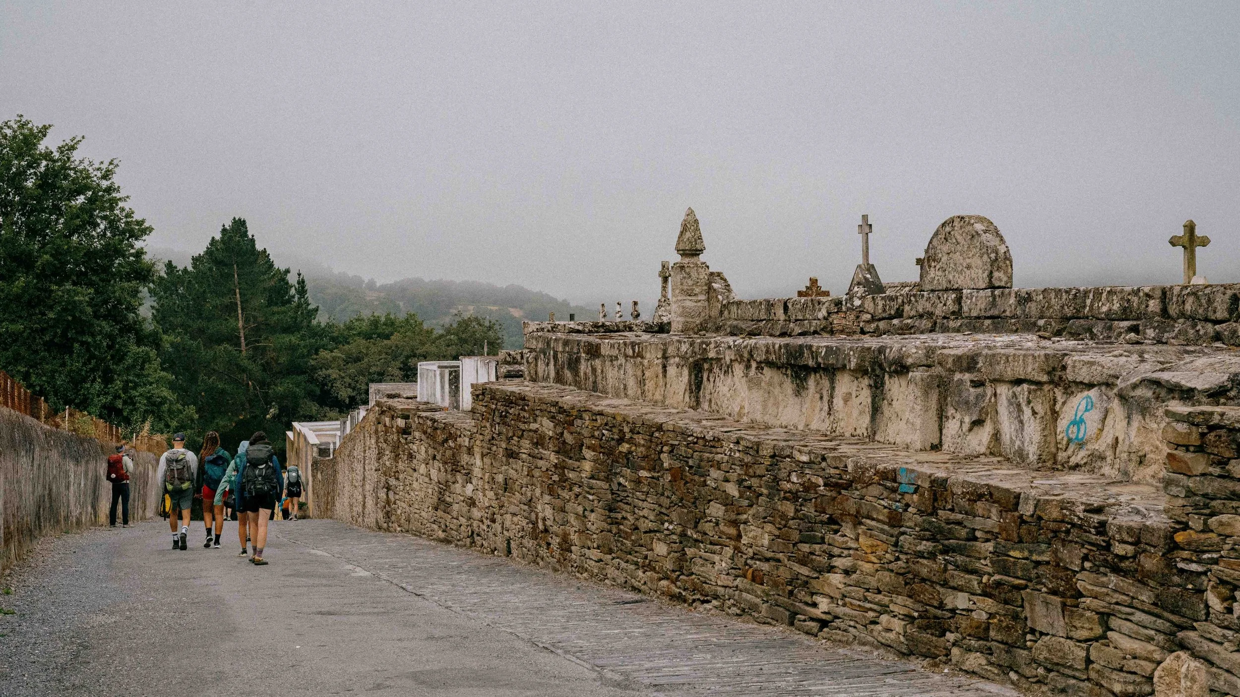 A group of hikers walking along a stone path beside an old stone wall with tombstones and crosses on top, surrounded by trees and overcast sky.