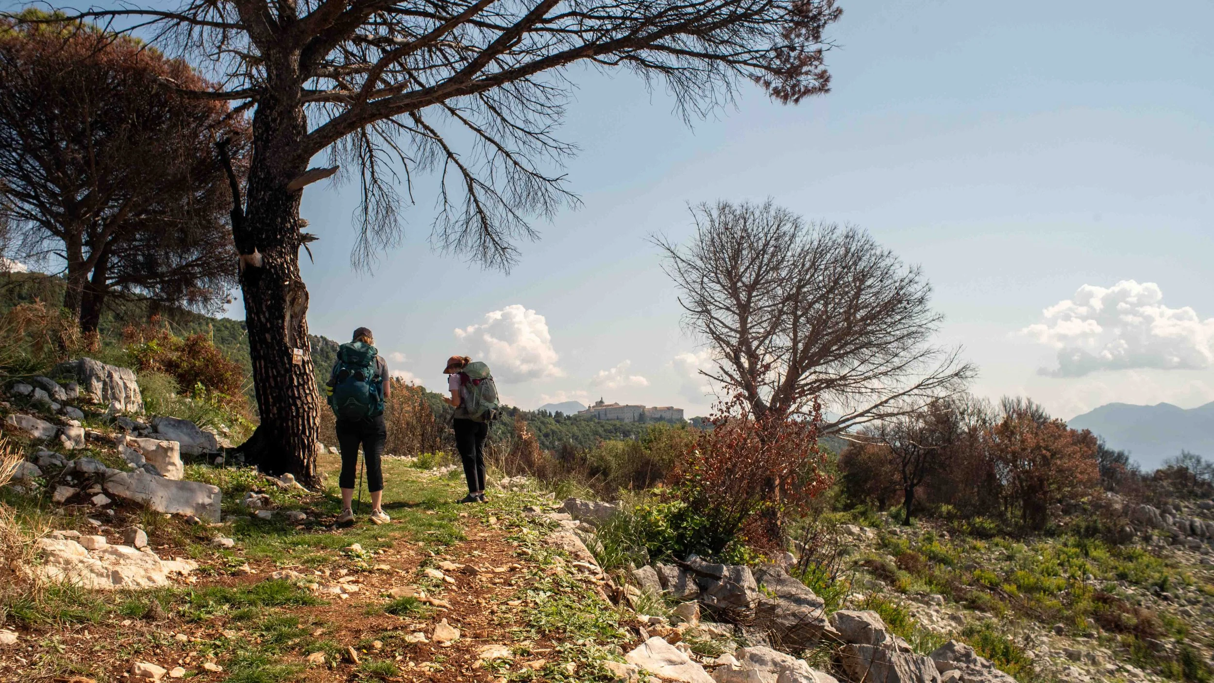 Two women hiking on a trail in a hilly area with trees and rocks, with mountains and a building in the distance.