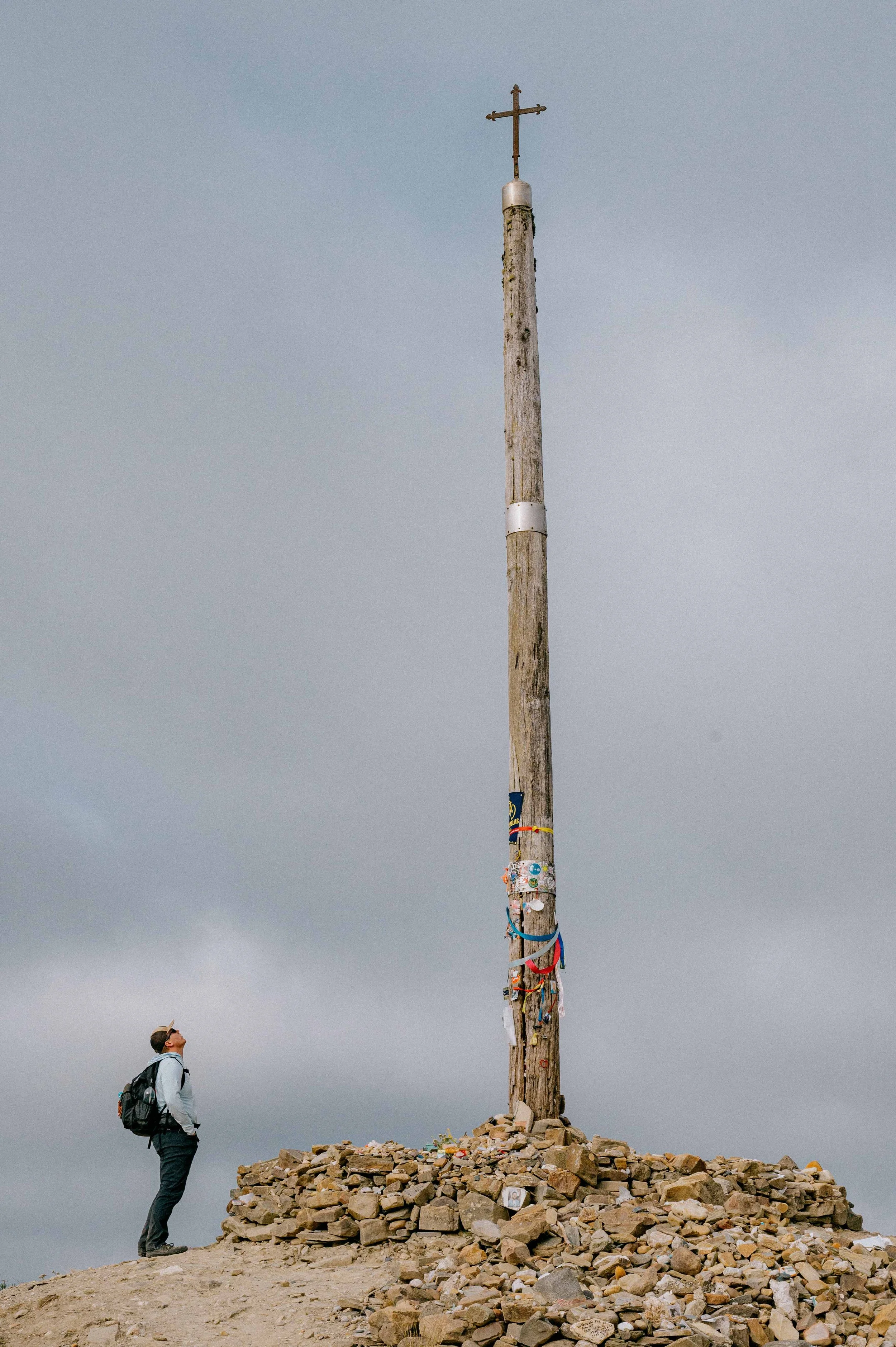 A person with a backpack looking up at a tall weathered wooden cross on a rocky mound, with a gray cloudy sky in the background.